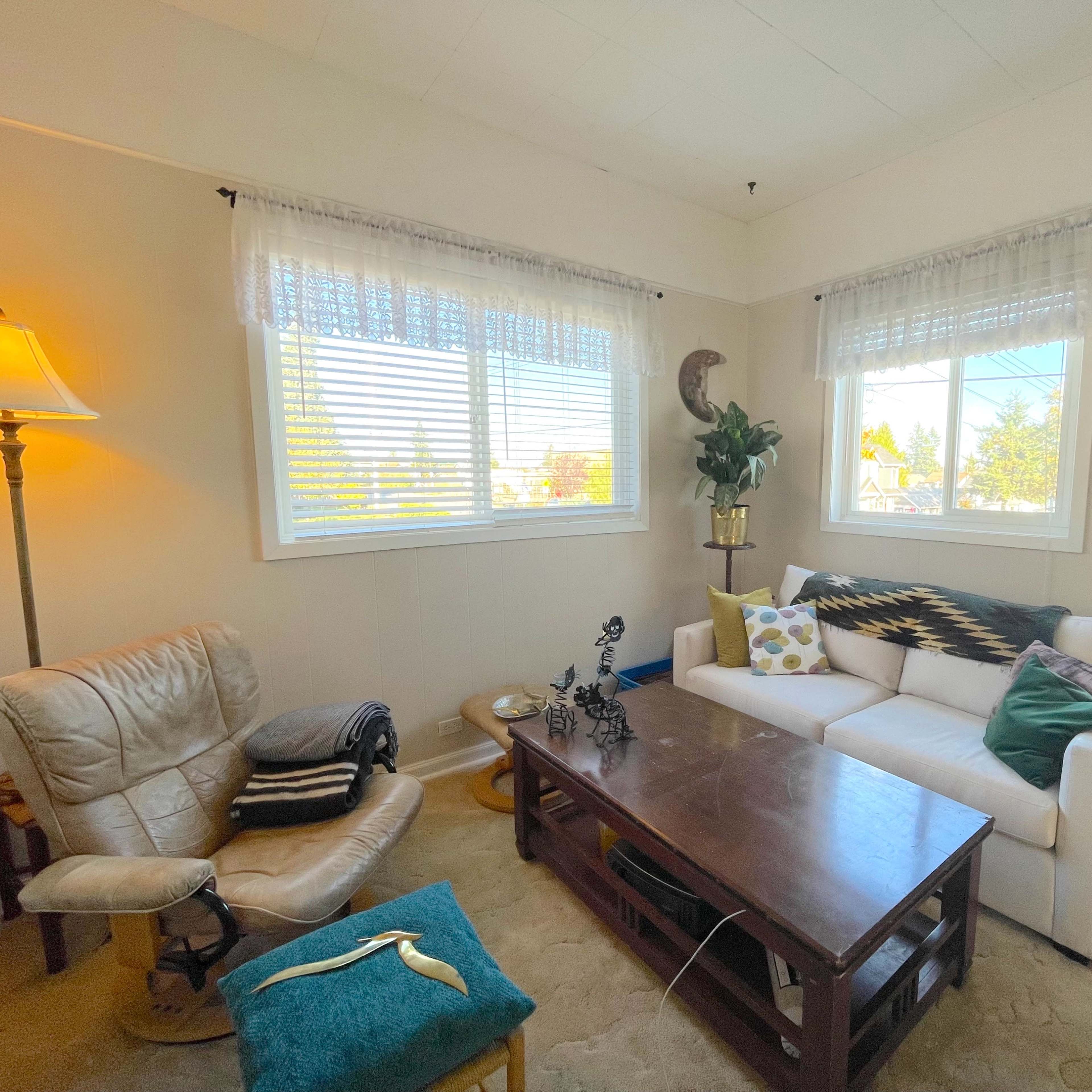 A cozy living room with a white sofa, a wooden coffee table, a comfortable armchair, and two windows allowing natural light to enter.