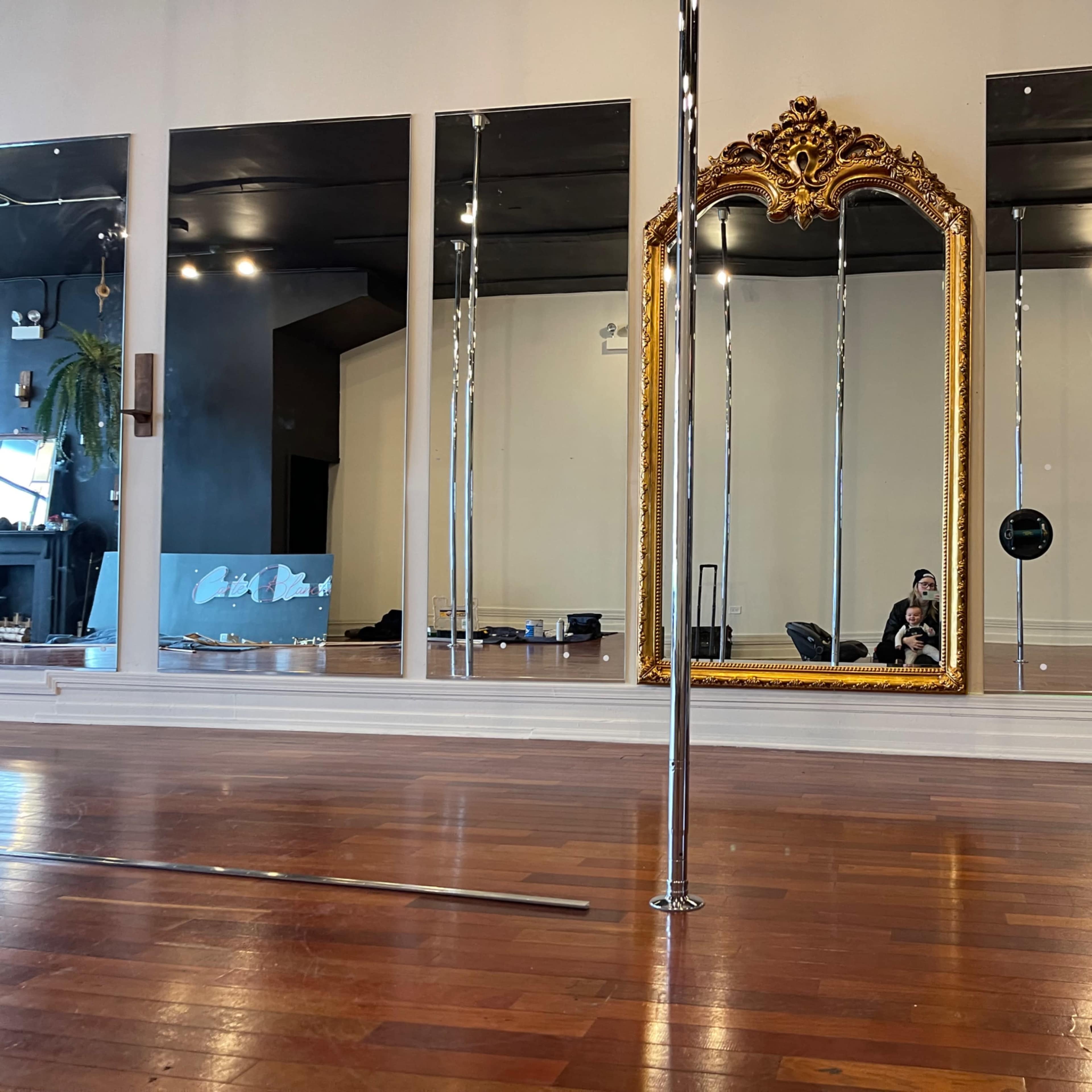 A spacious room with polished wooden floors, featuring large mirrors, a decorative golden mirror, and a pole in the center.