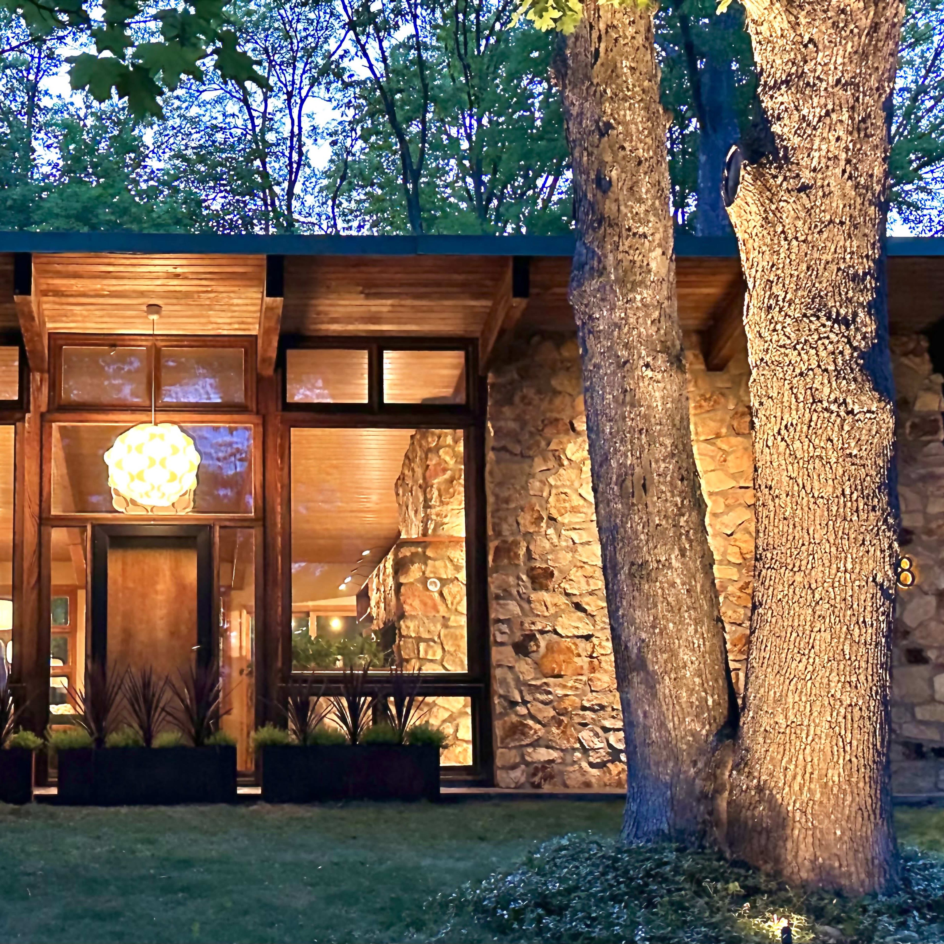 A modern stone and wood house features large windows and a distinctive hanging light fixture, set amidst trees in a landscaped yard.