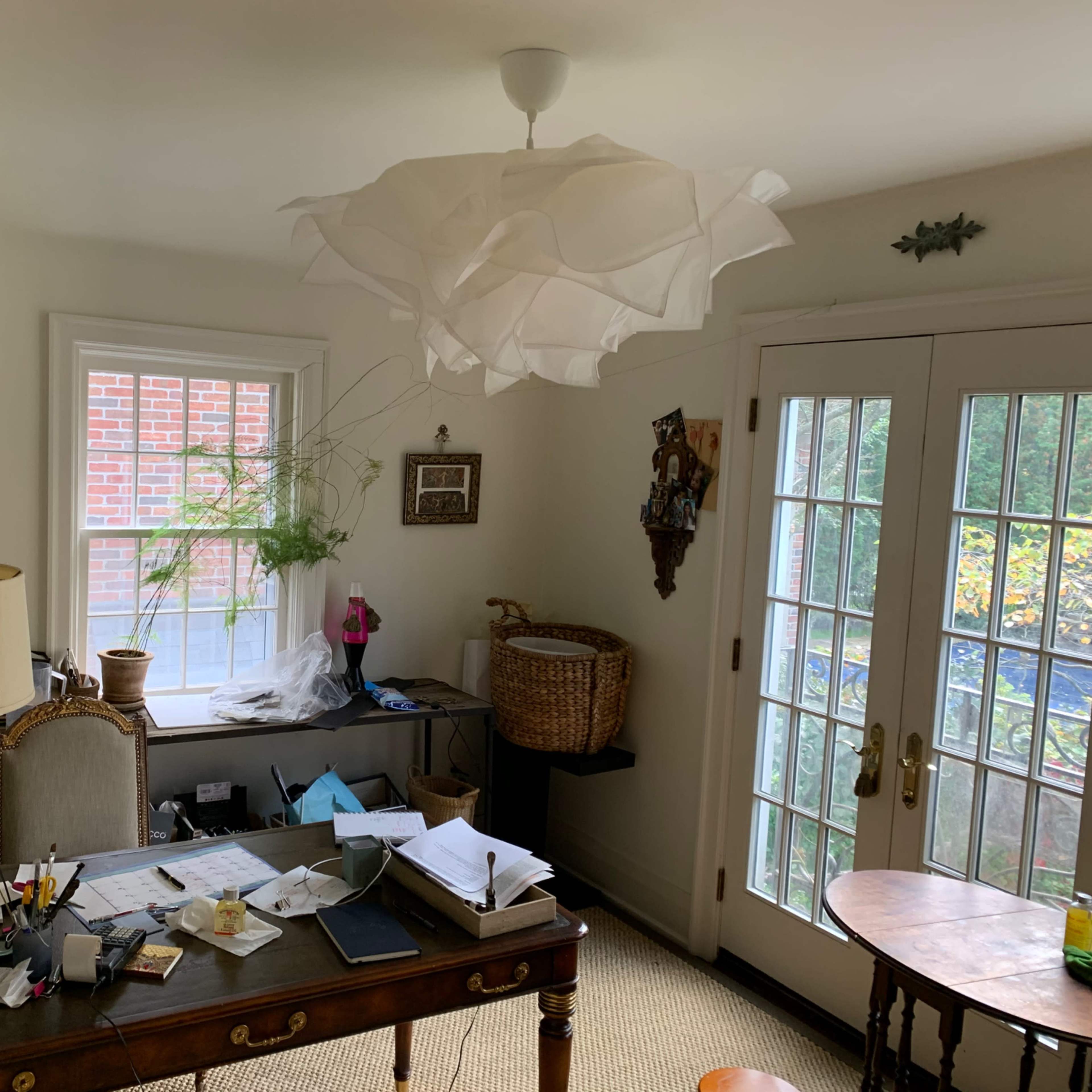 The room features a desk cluttered with papers and a lamp, a large window with plants, and a door leading outside.