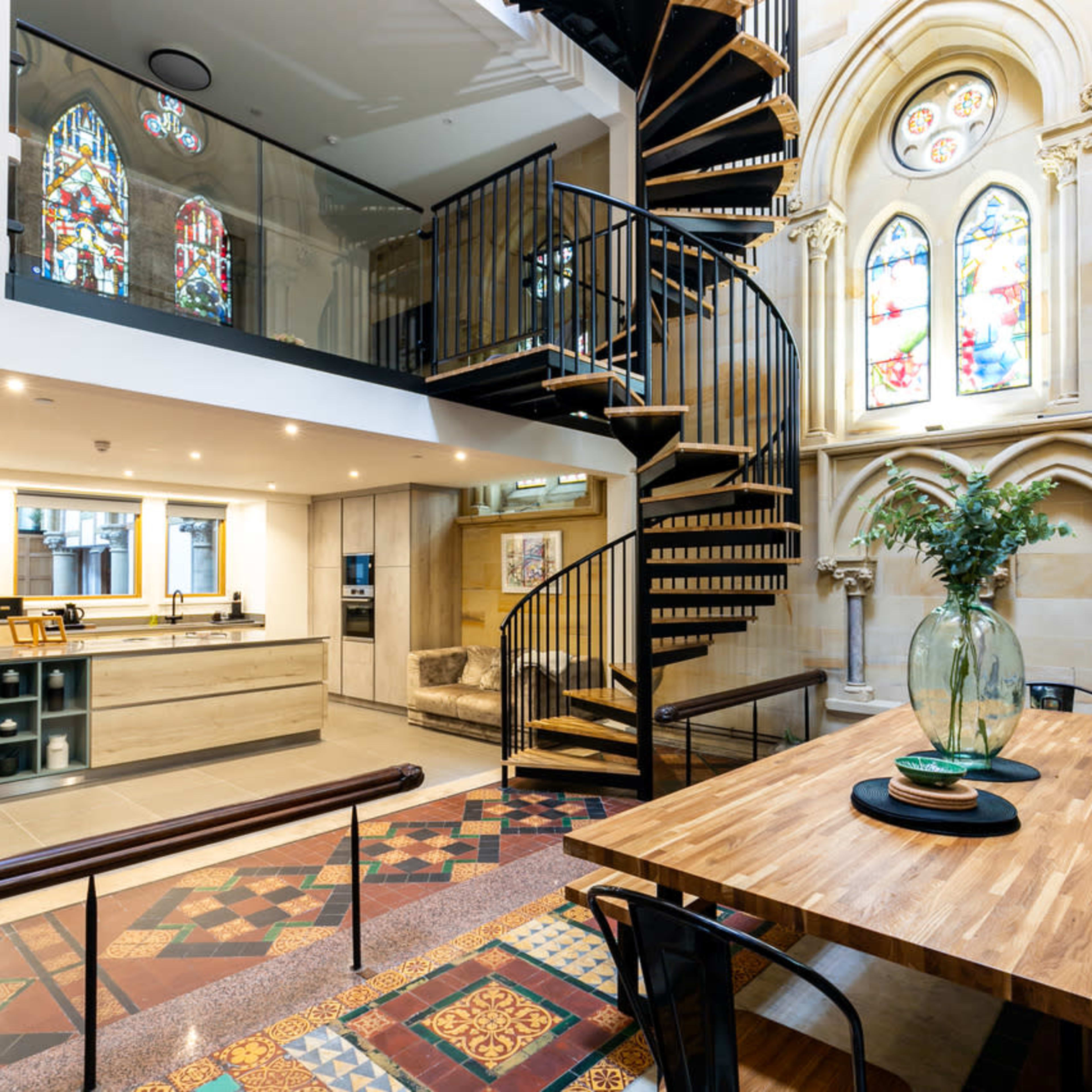 A modern interior space featuring a spiral staircase, a kitchen area, and a dining table surrounded by colorful tiled flooring and large stained glass windows.