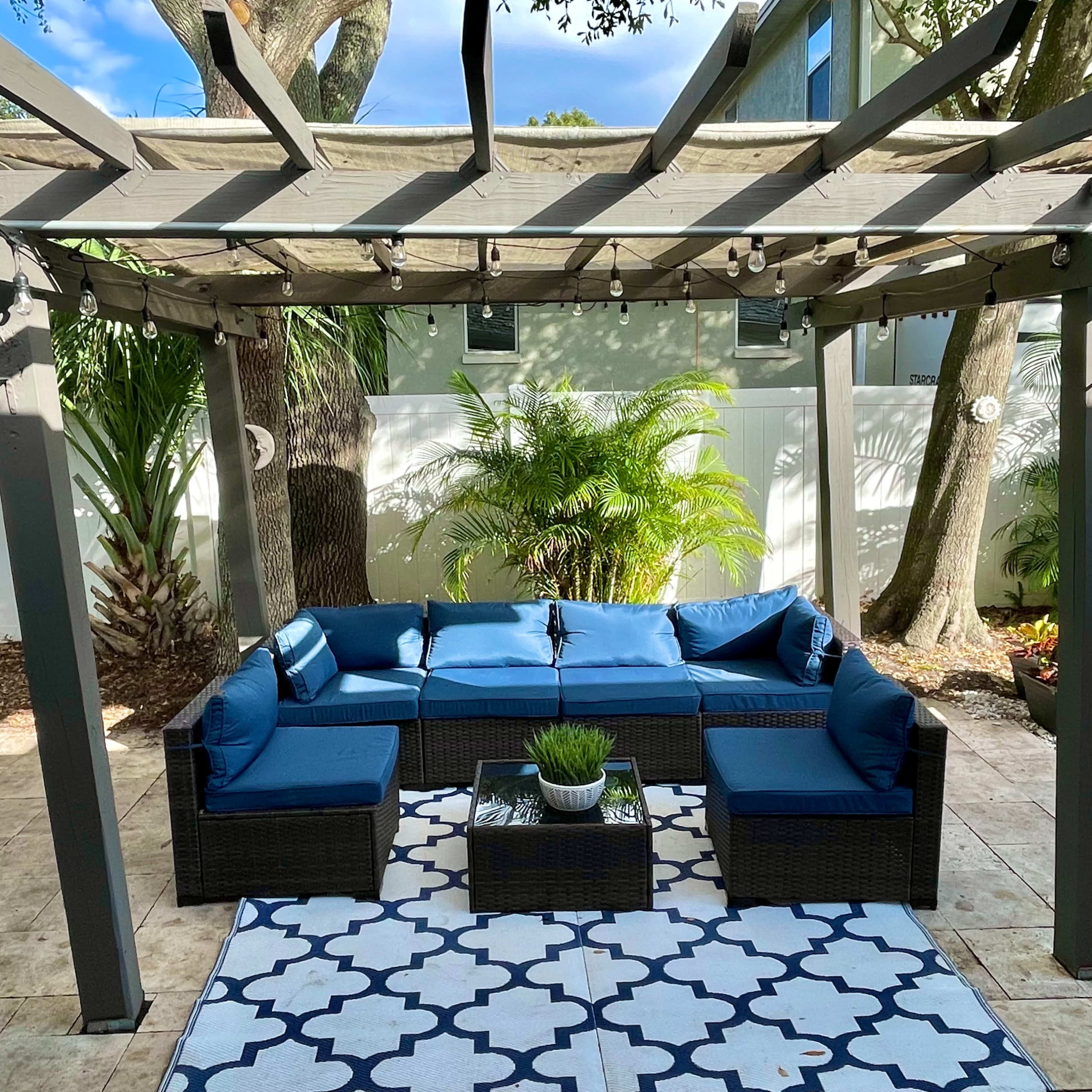 A shaded outdoor seating area features a sectional sofa with blue cushions, a coffee table, and a patterned rug surrounded by greenery and a trellis.