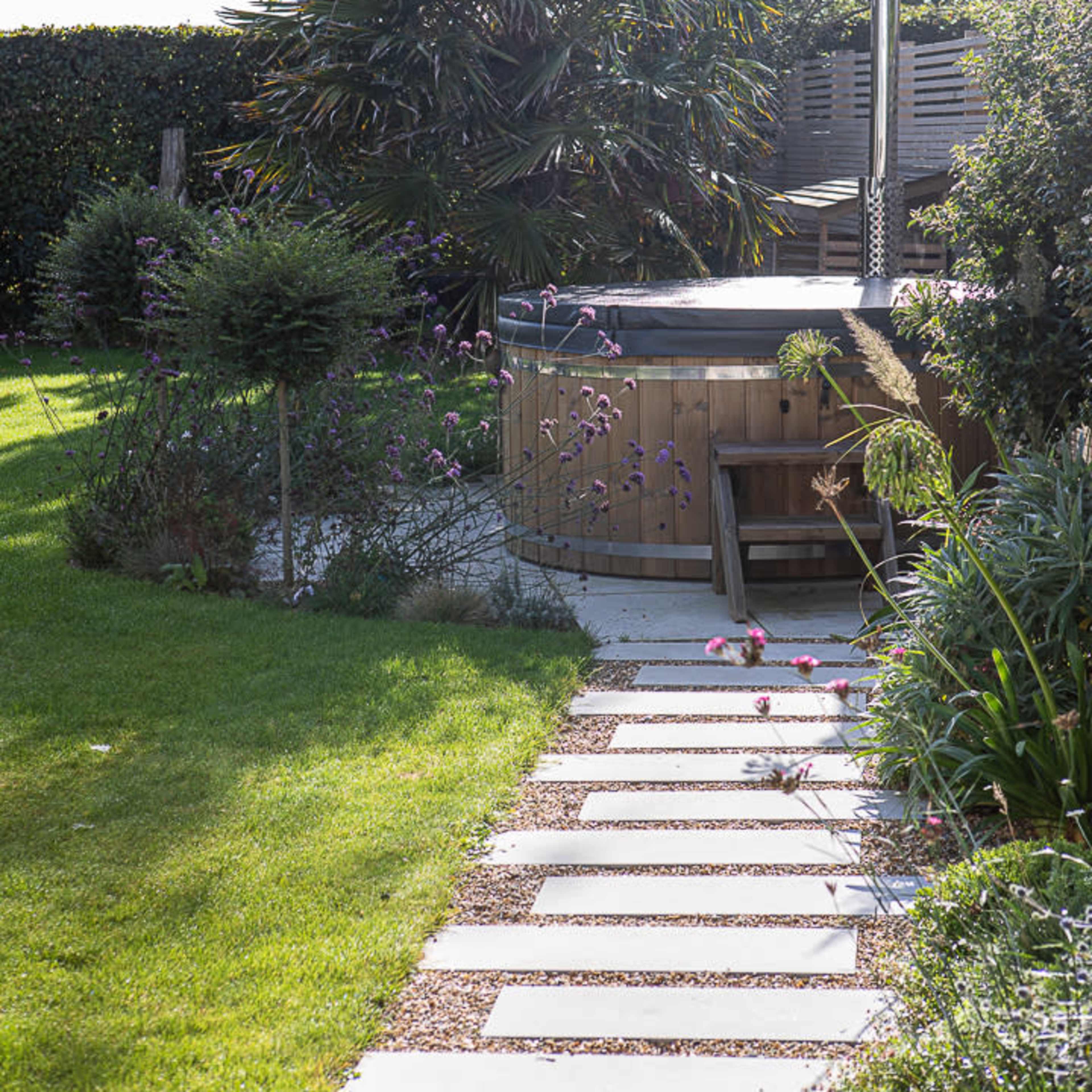 A landscaped garden with a stone pathway leading to a wooden hot tub surrounded by greenery and flowers.