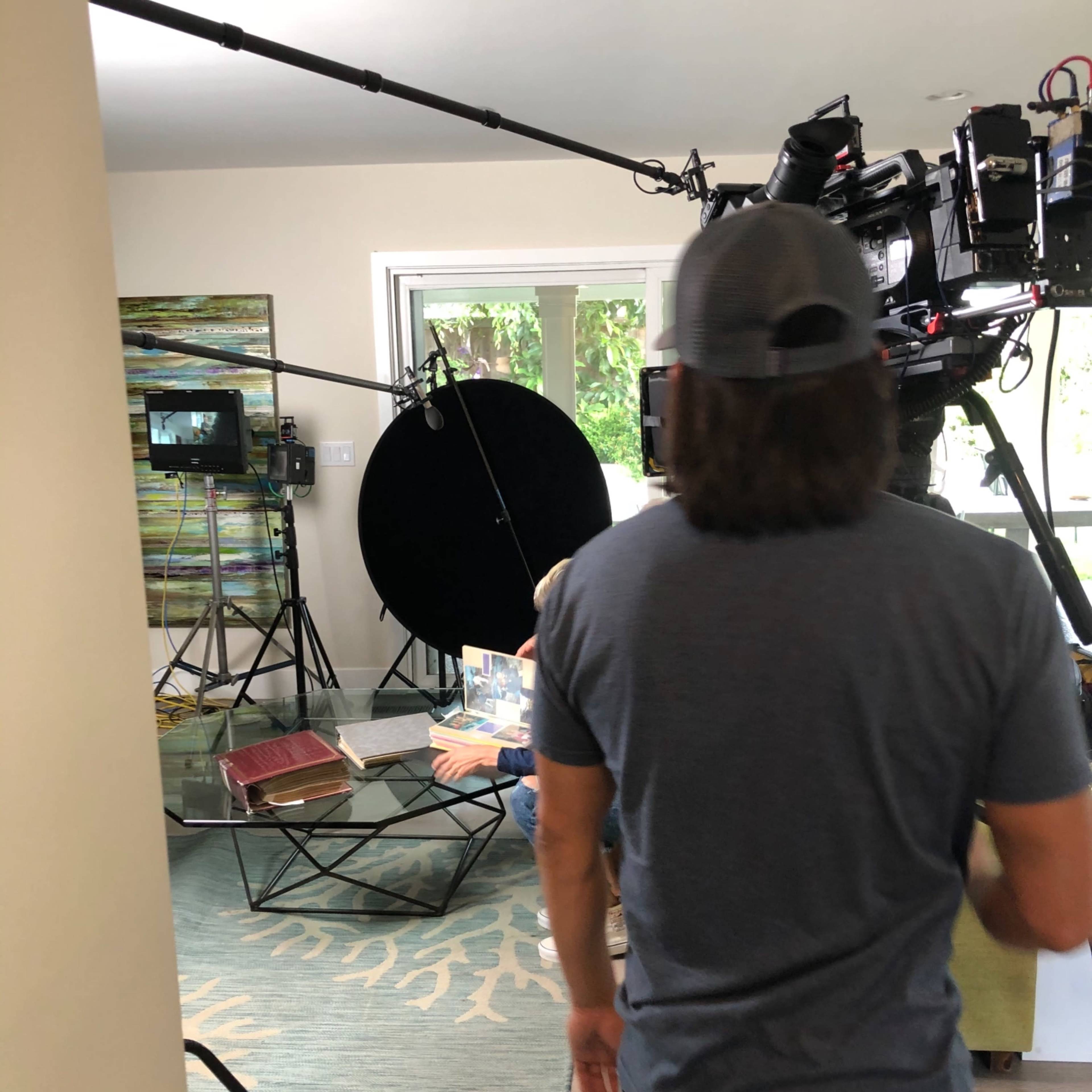 A crew member stands in a living room set up for filming, surrounded by cameras, lights, and a reflector.