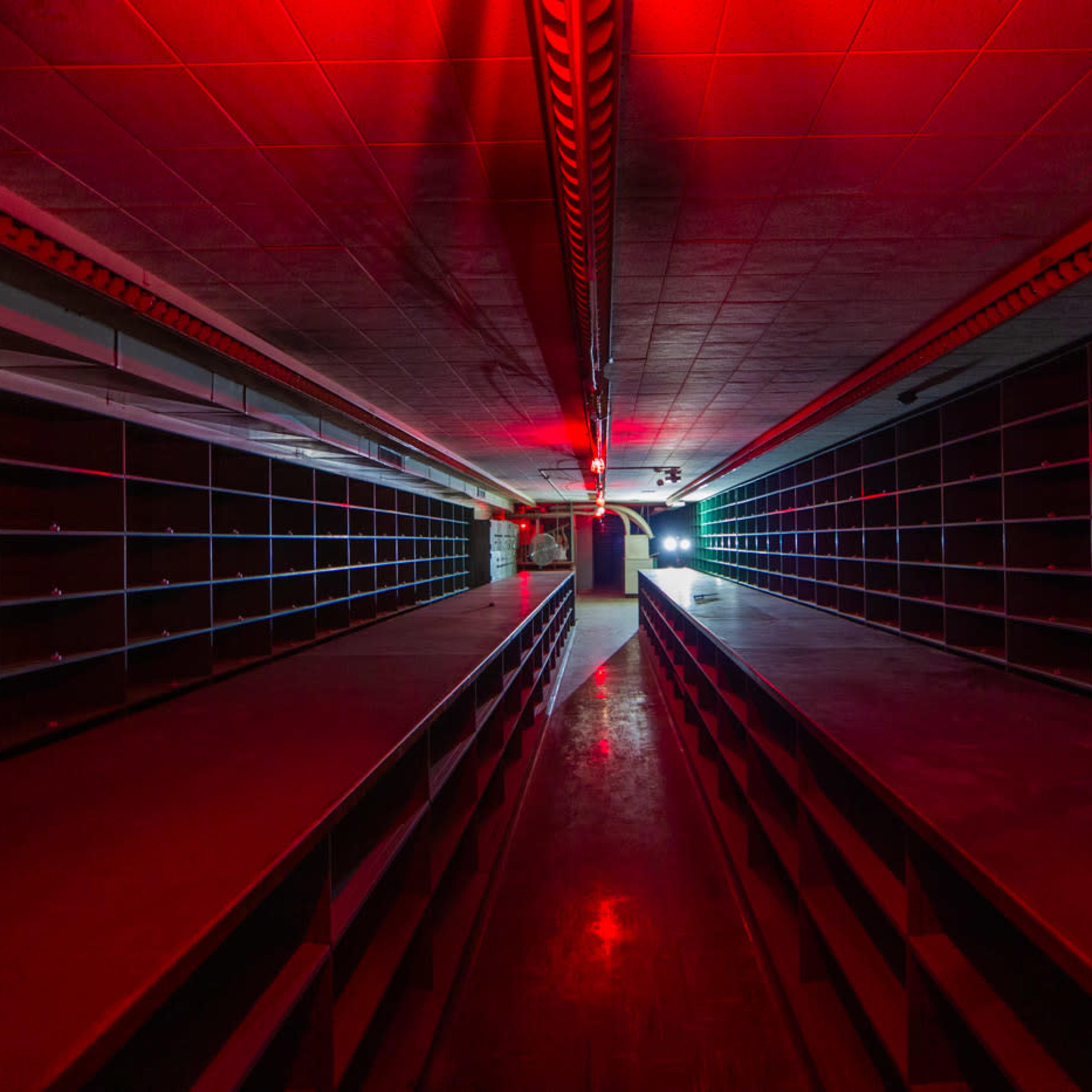 A dimly lit storage room features long shelves lined along both sides, illuminated by red and blue lights at either end.