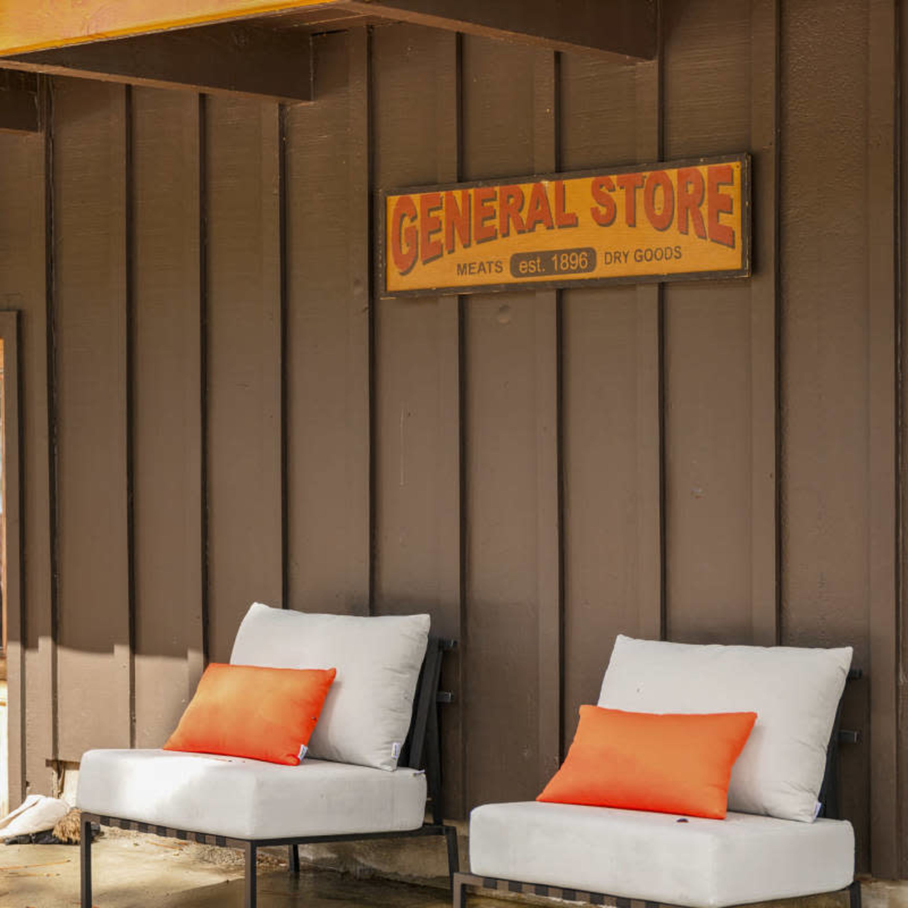 Two white chairs with orange pillows positioned under a sign that reads "GENERAL STORE" on a brown wooden wall.