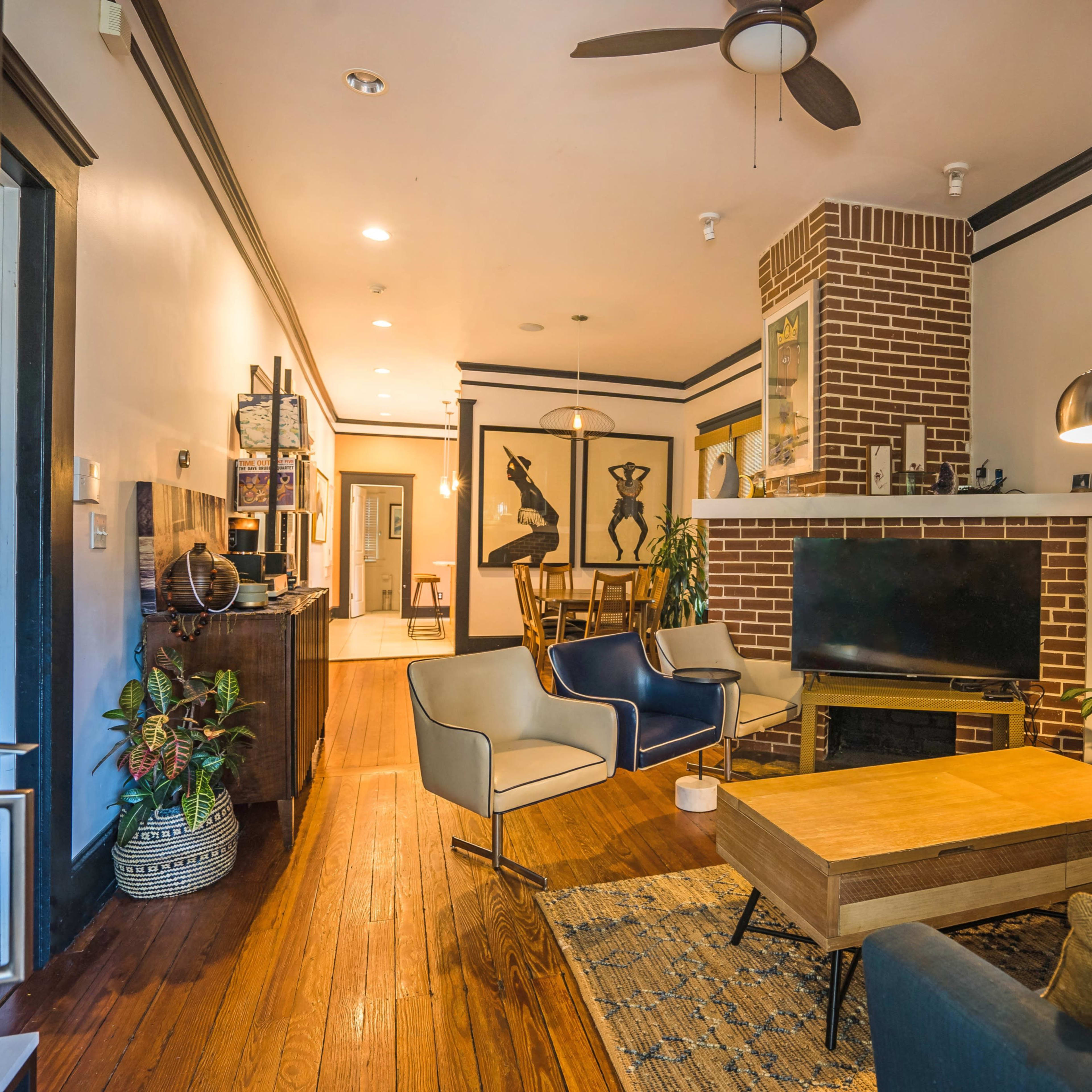 A cozy living room featuring wooden floors, a brick fireplace, modern furniture, and various plants.