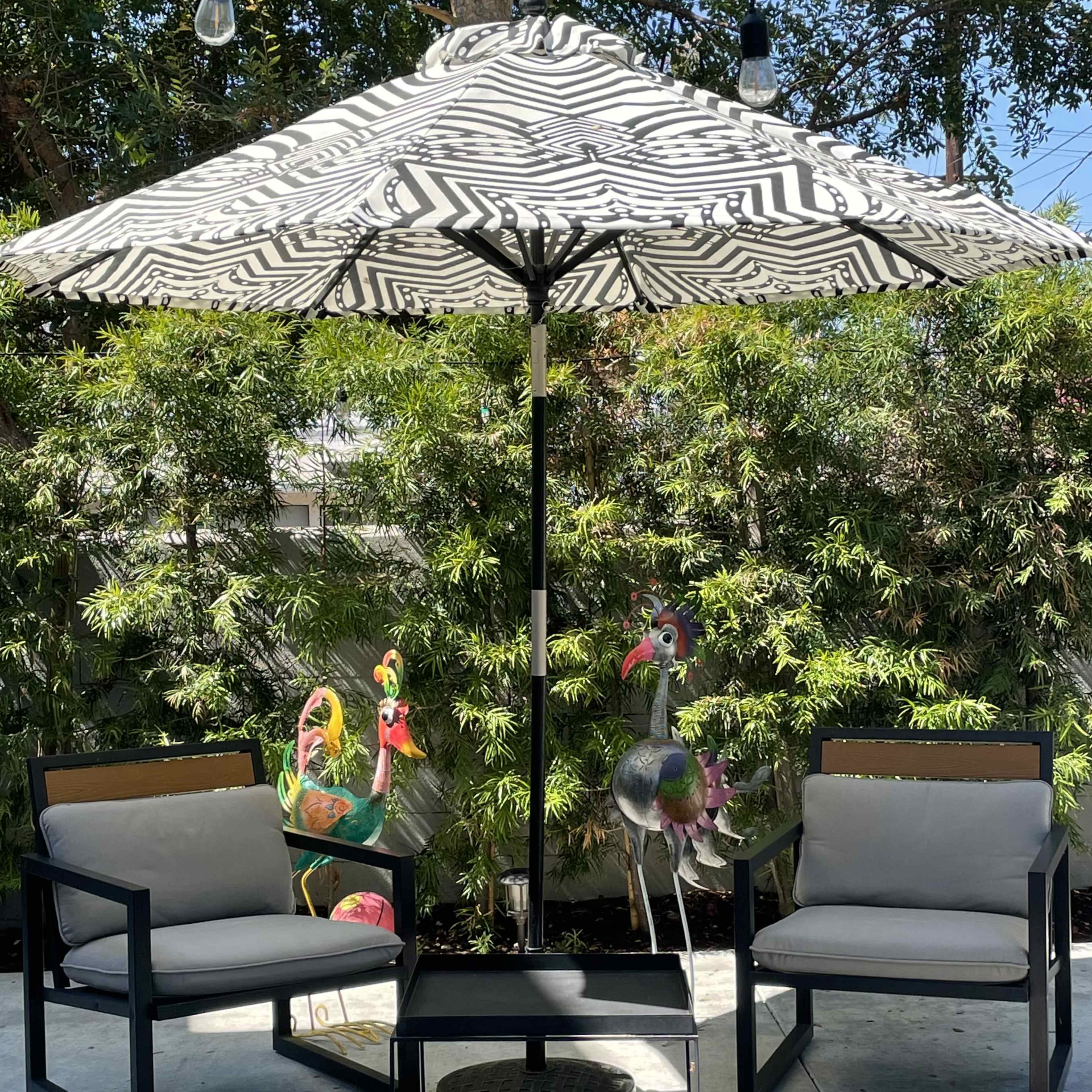 A patio area features two black chairs and a table under a large striped umbrella, surrounded by bamboo plants and decorative flamingo statues.