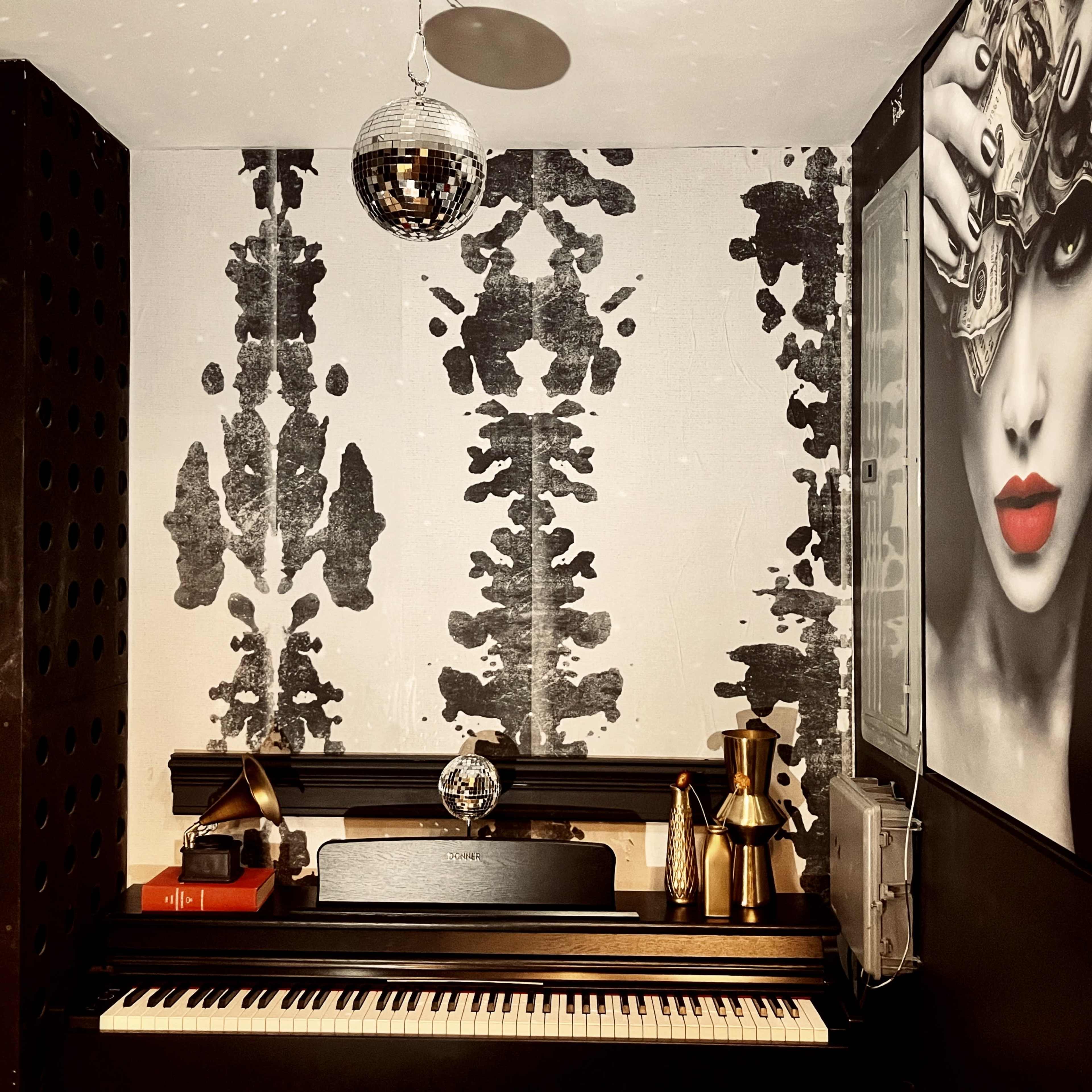A black piano placed against a patterned wall, adorned with a disco ball and a framed portrait of a woman with striking red lips.