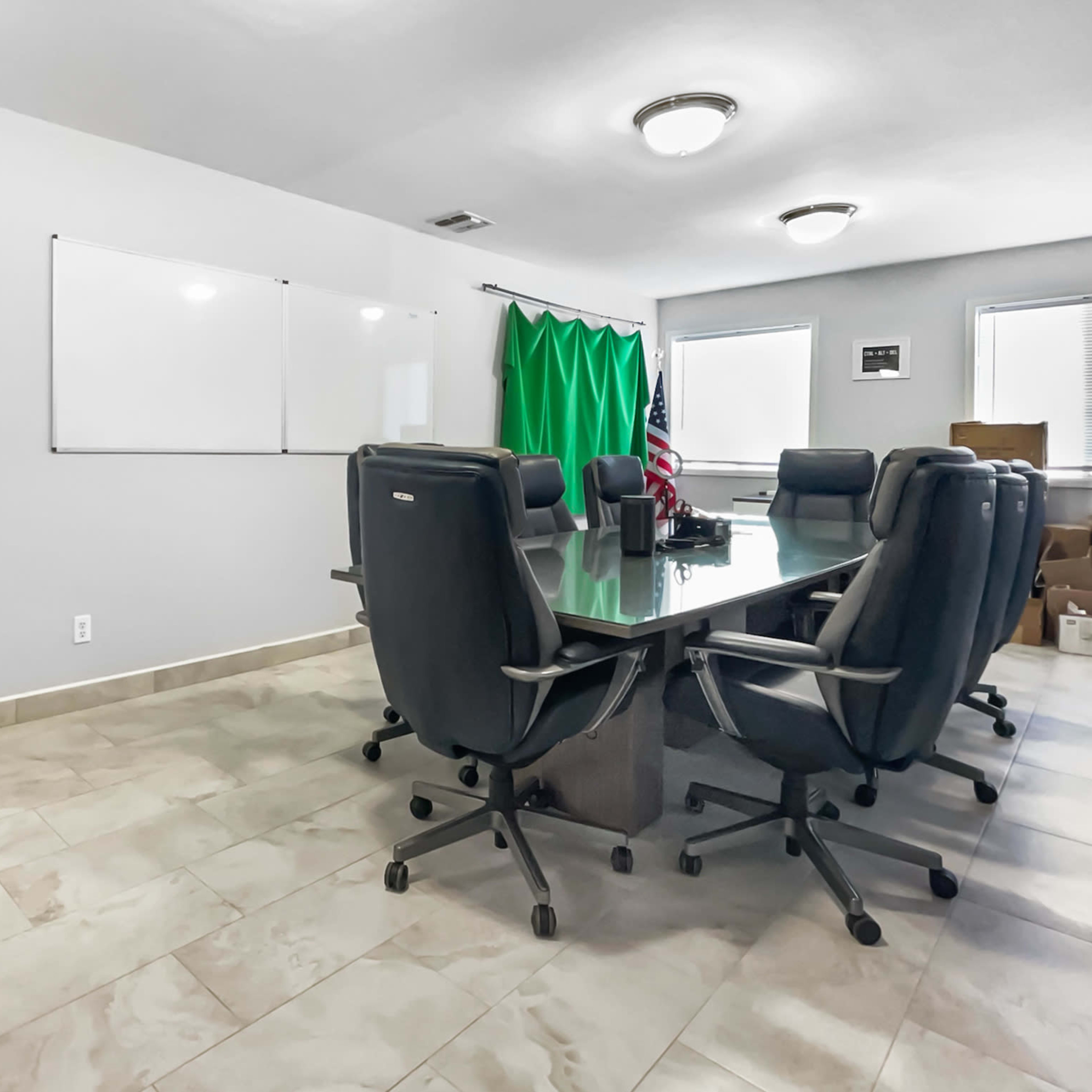 A meeting room features a large glass table surrounded by black office chairs, with a whiteboard and a green backdrop on the walls.