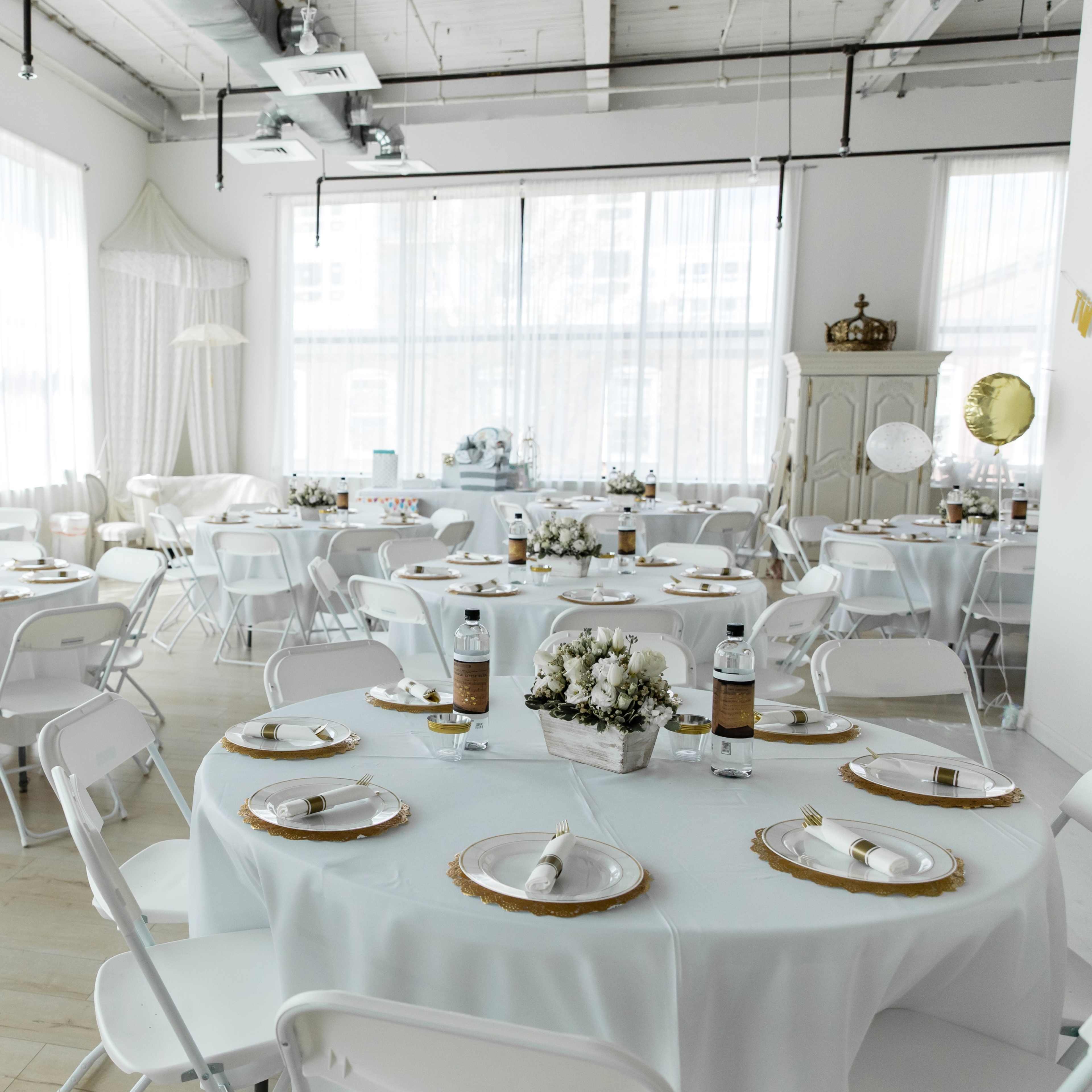 A bright event space with multiple round tables set for a gathering, each adorned with white tablecloths and decorative centerpieces.