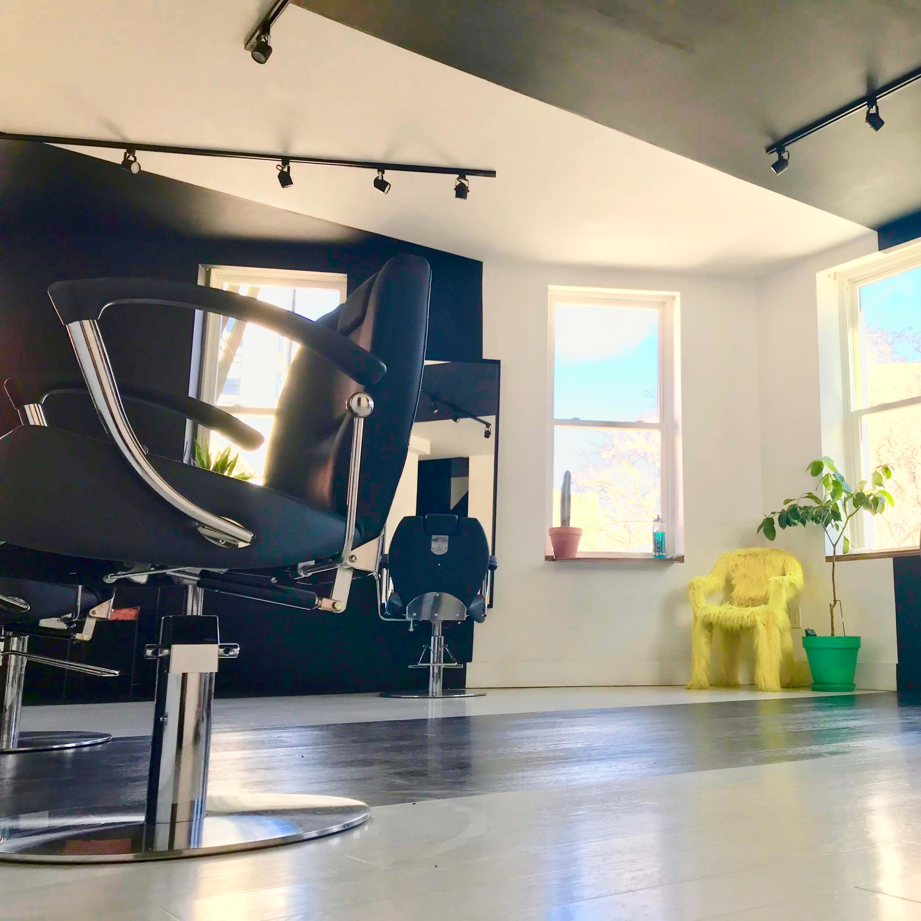 A modern hair salon with several black styling chairs, large windows, and a yellow armchair in the corner.