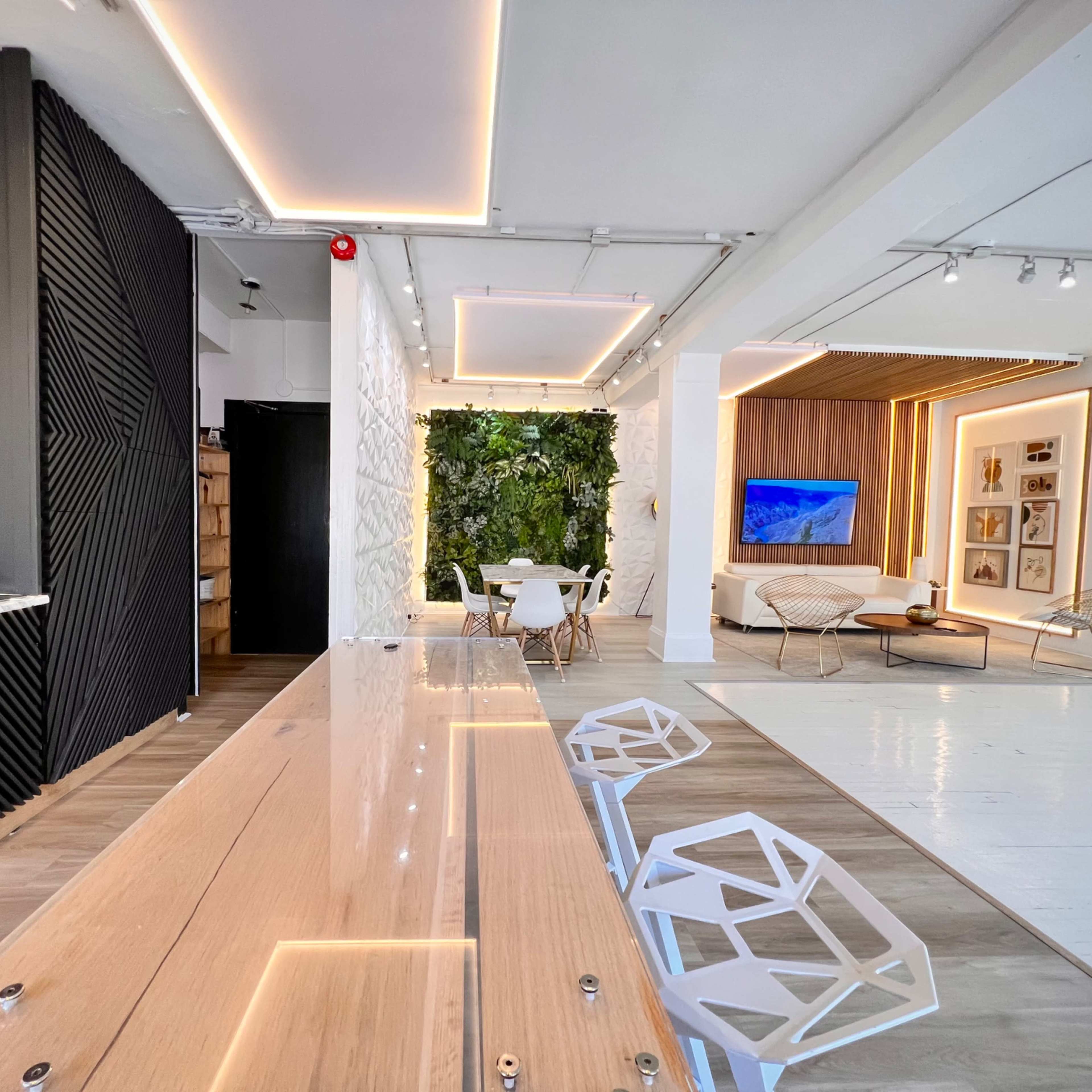 A modern interior space featuring a kitchen area with a long wooden table and white chairs, leading to a lounge area with a television and greenery.