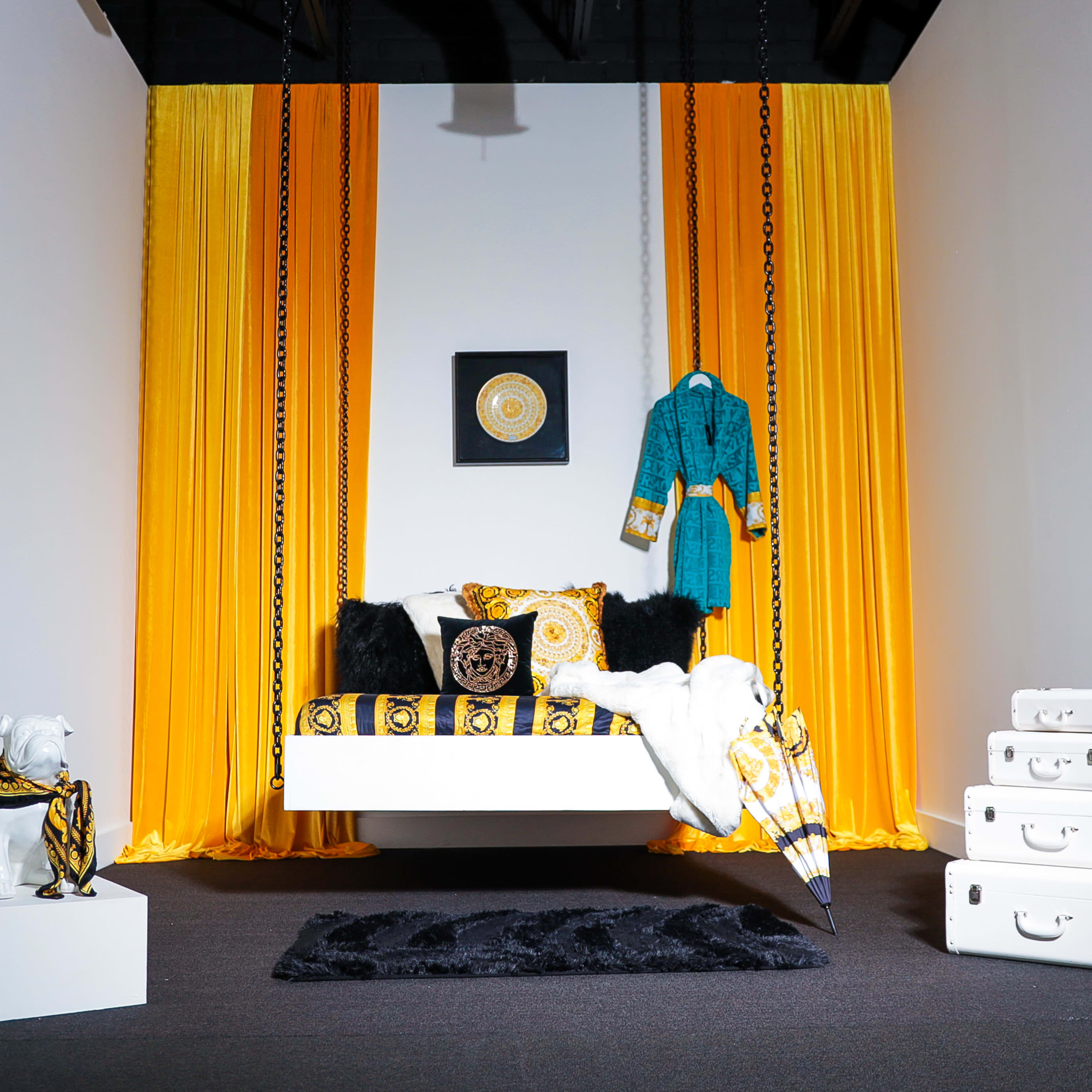 A modern room features a suspended bed with yellow and black patterned bedding, a decorative robe hanging from the ceiling, and white luggage stacked beside a small sculpture.