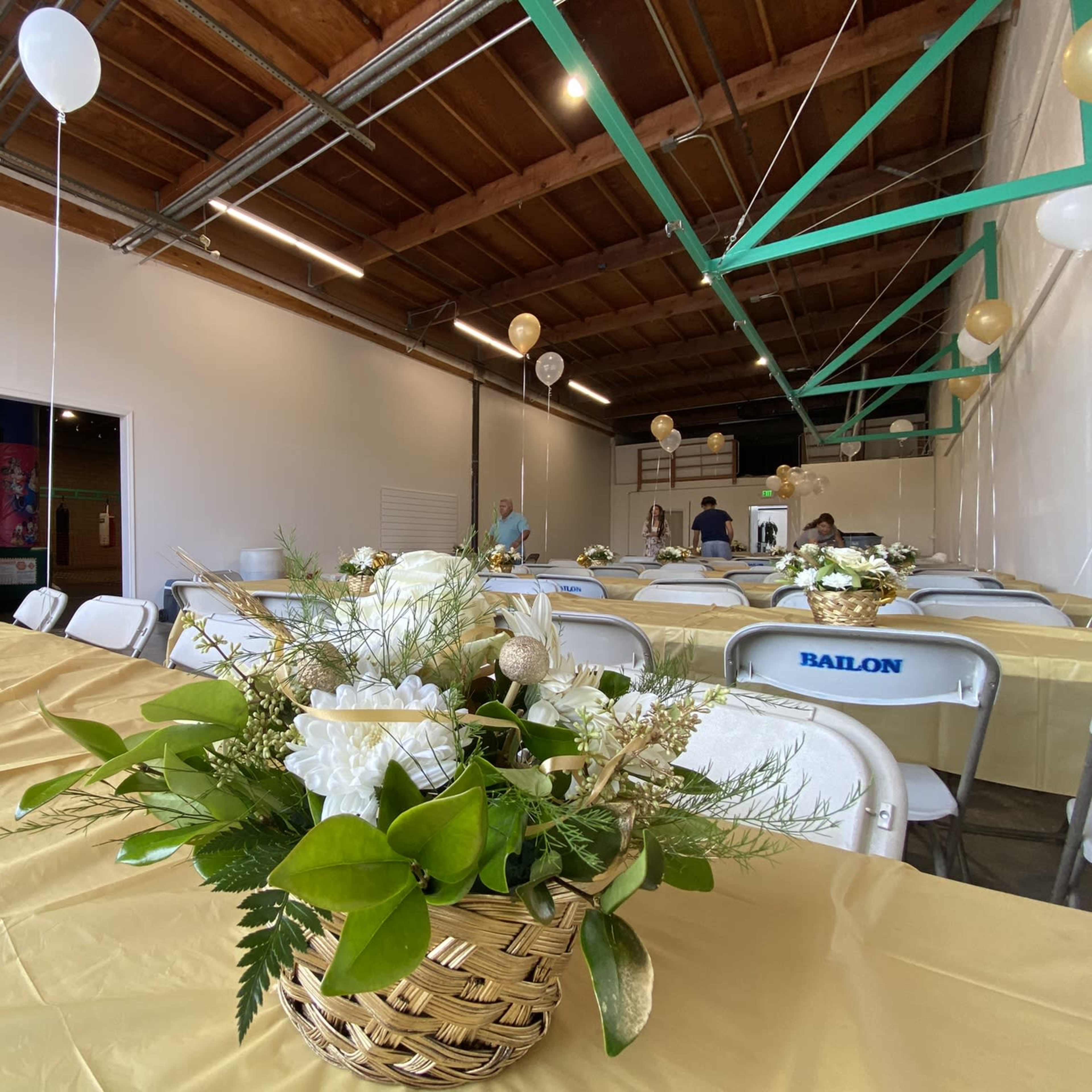 A spacious room with tables set for an event, adorned with floral centerpieces and balloons hanging from the ceiling.