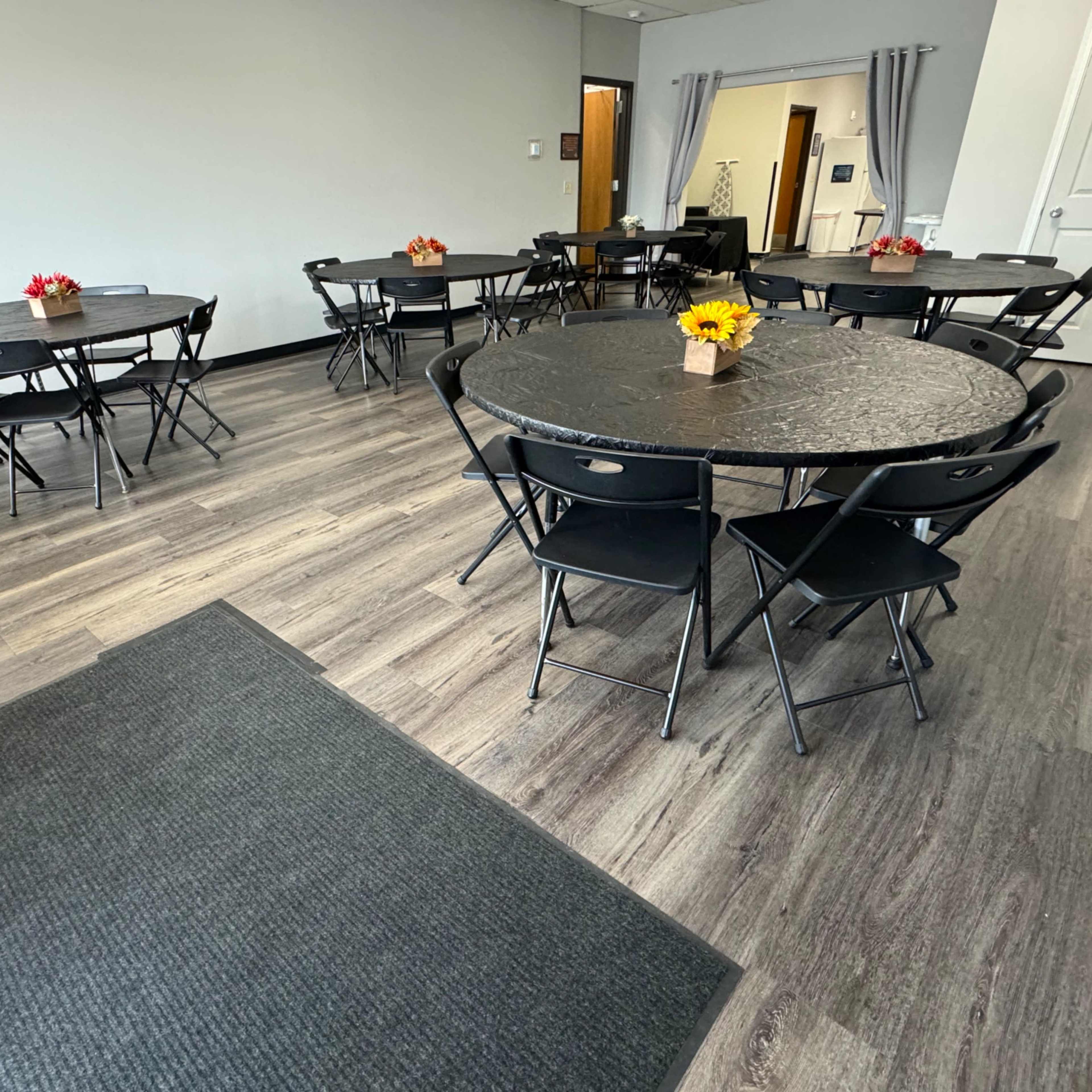 A well-lit meeting room with several round black tables set with small flower centerpieces, arranged on a wooden floor.