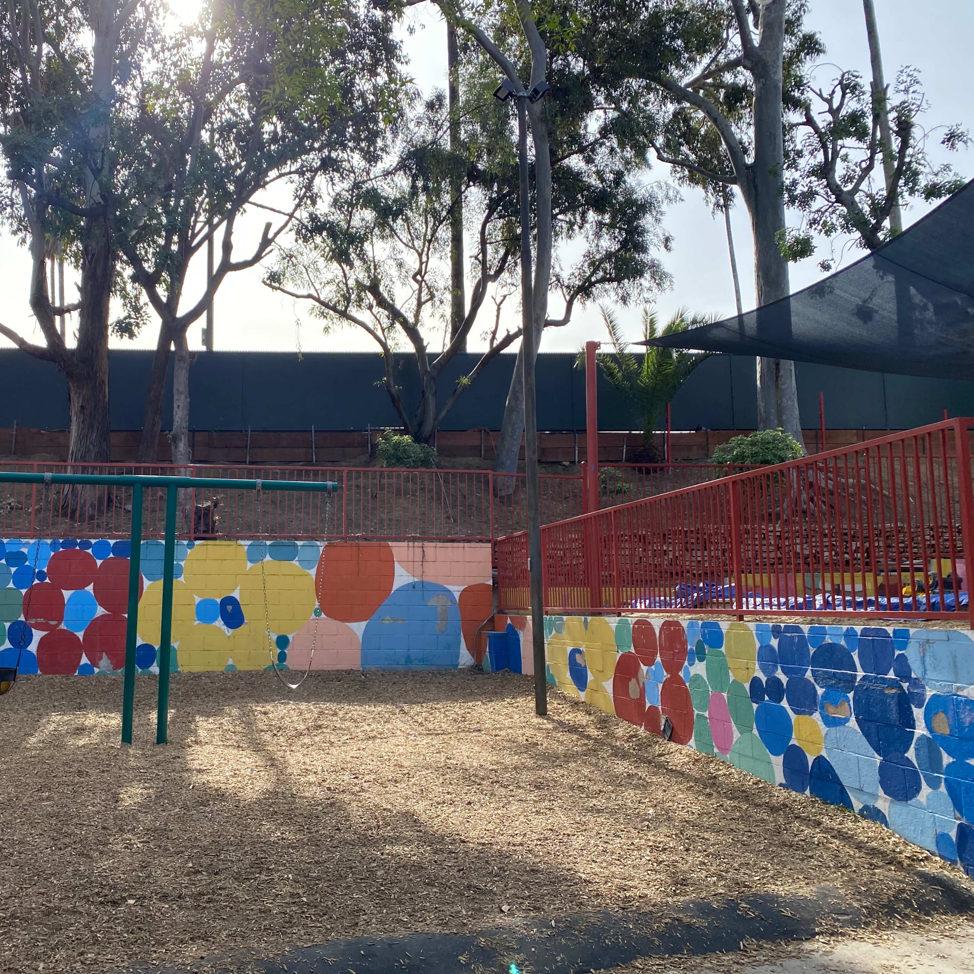 A playground area with colorful murals on the walls, a swingset, and wood chips covering the ground under trees.