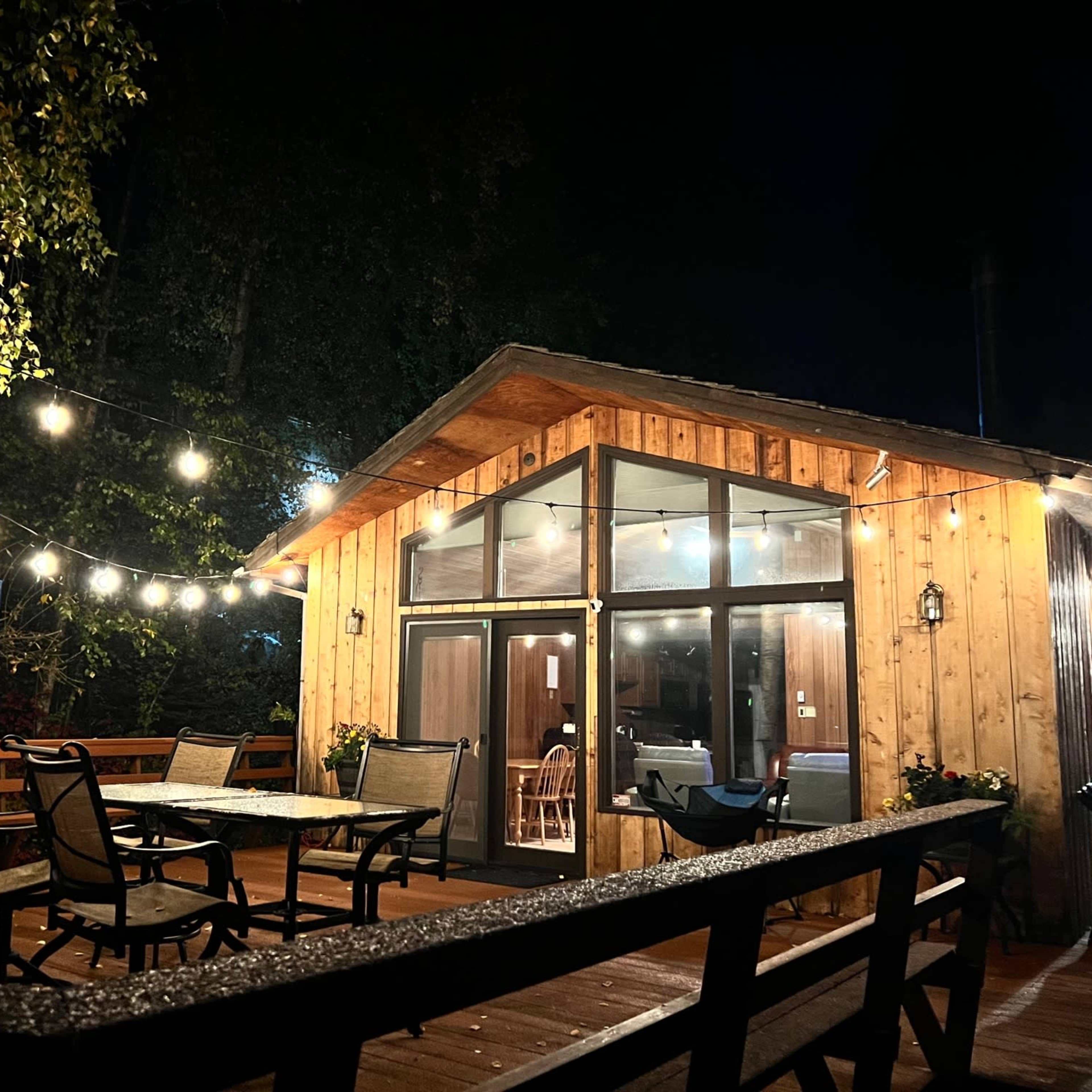A wooden cabin with large windows is illuminated by string lights in the evening.