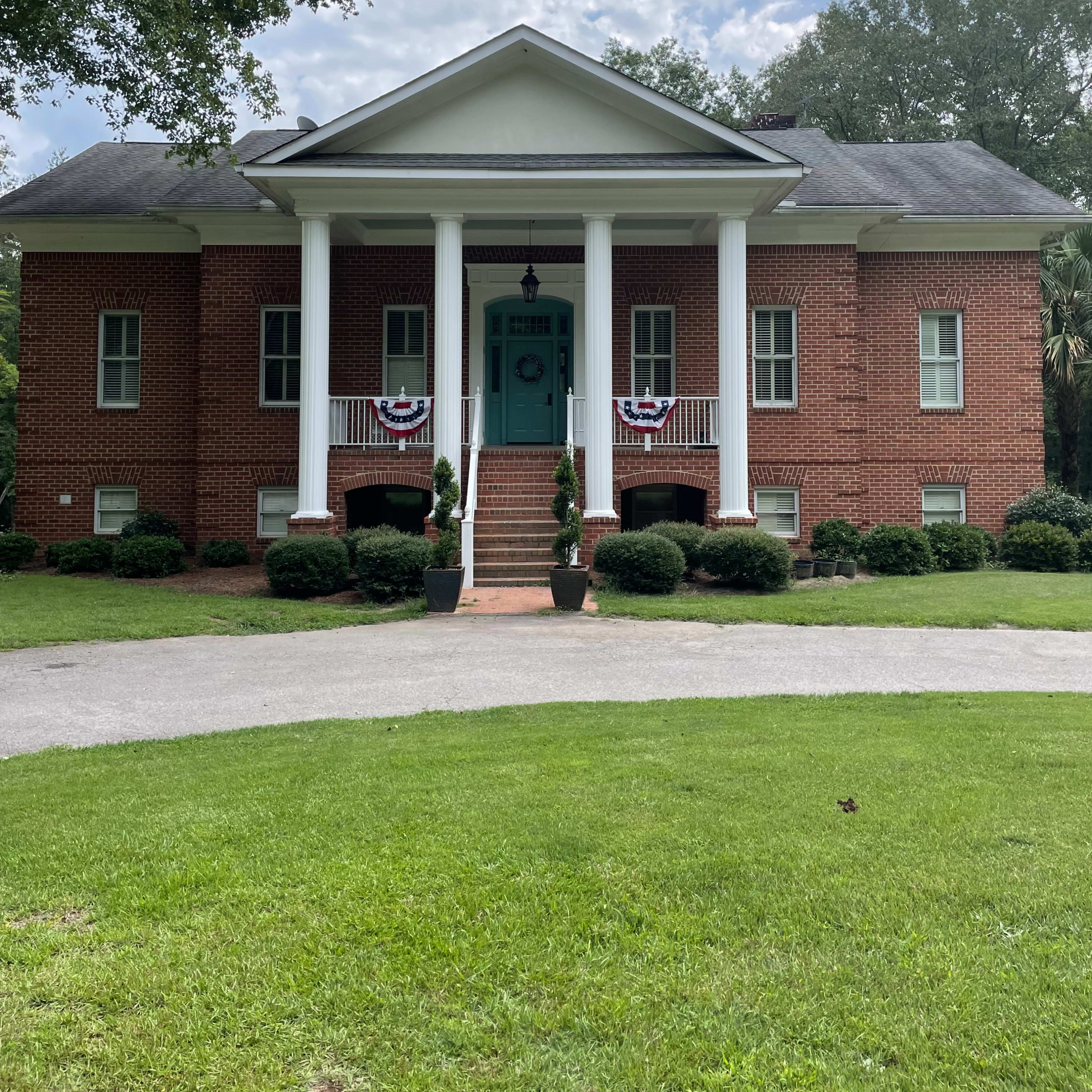 A two-story brick house with white columns, a green door, and decorative bunting on the front porch is set on a grassy lawn.