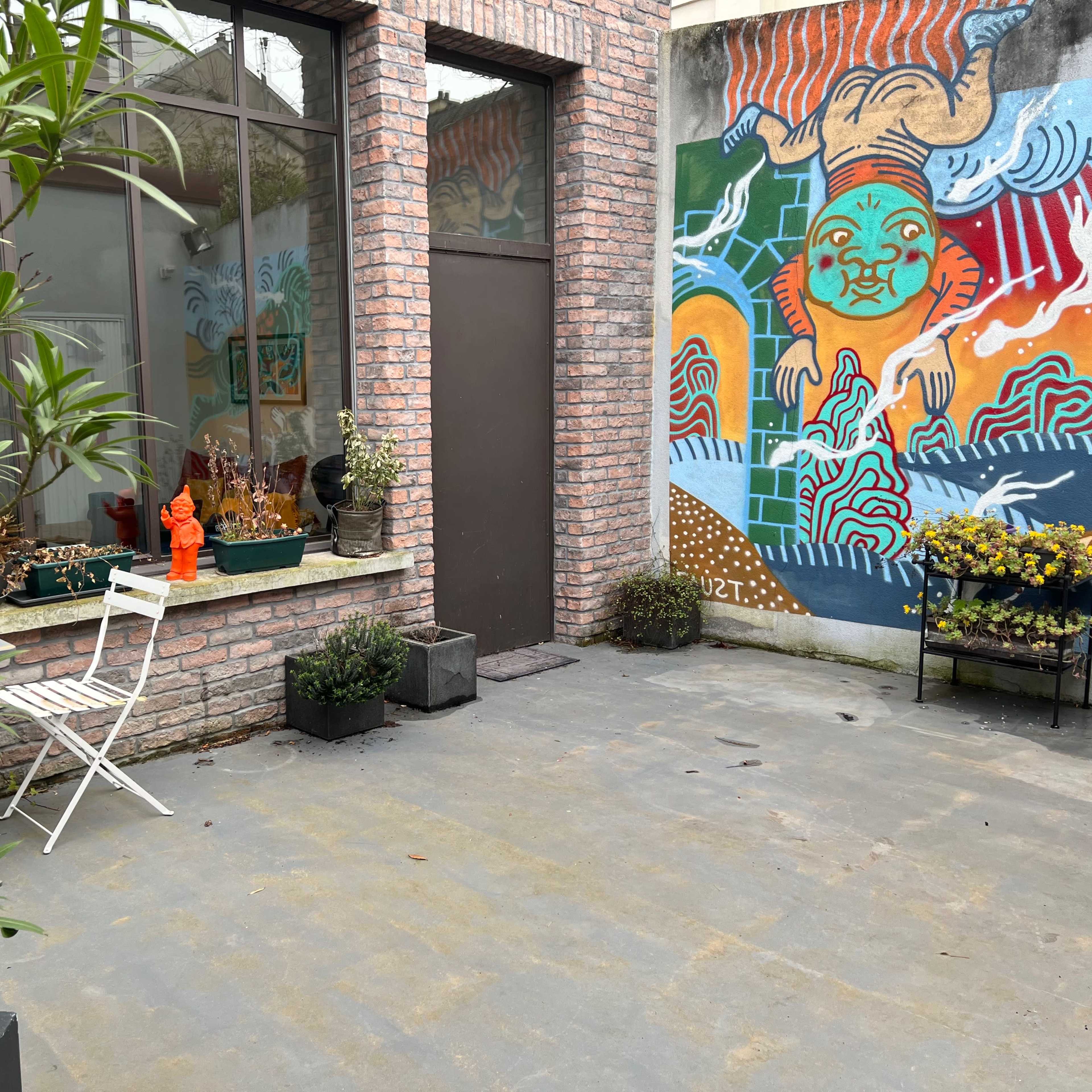 A small outdoor space featuring potted plants, a white metal chair, and a colorful mural on a brick wall.