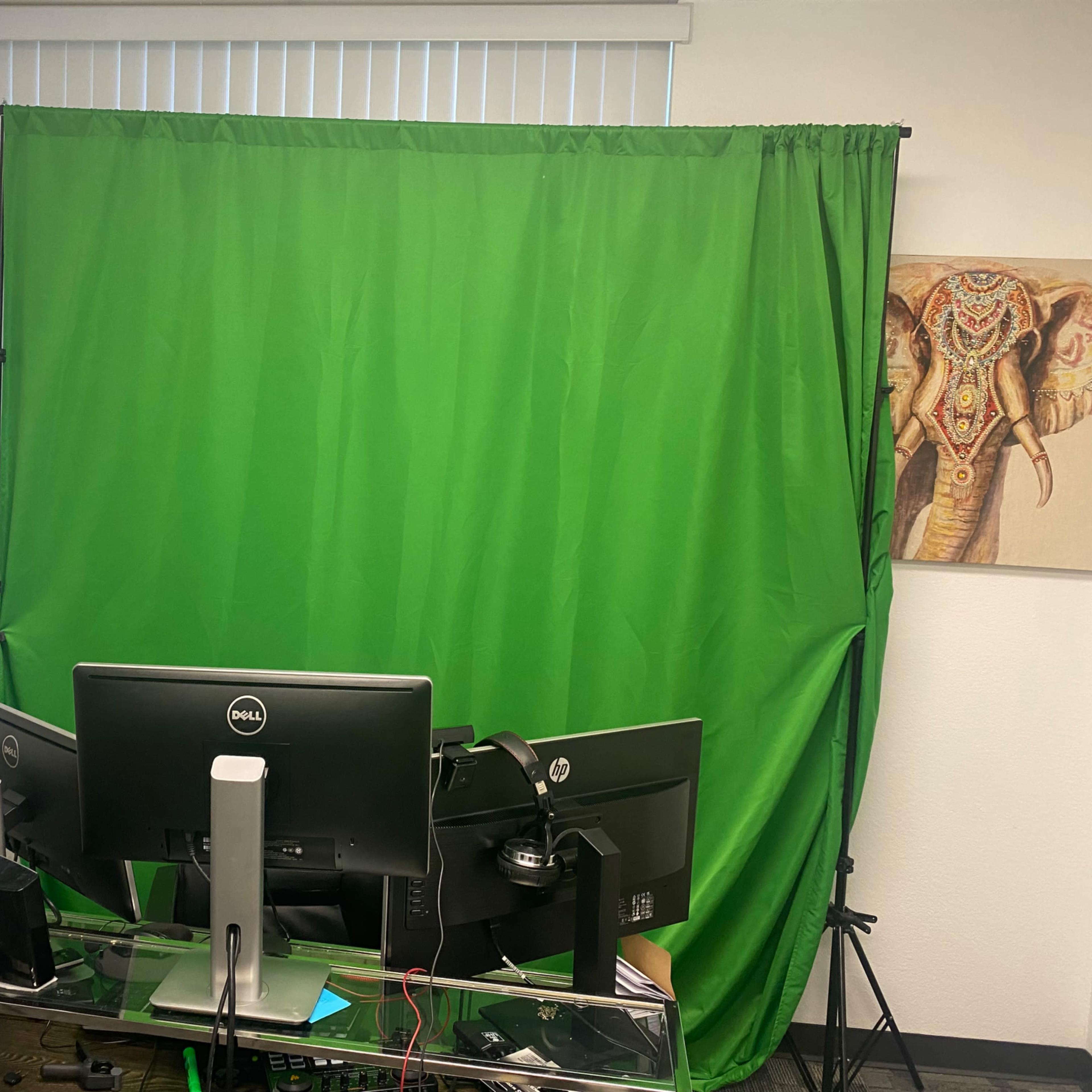 A workspace featuring a green screen setup behind dual computer monitors, with a decorative elephant painting on the wall.