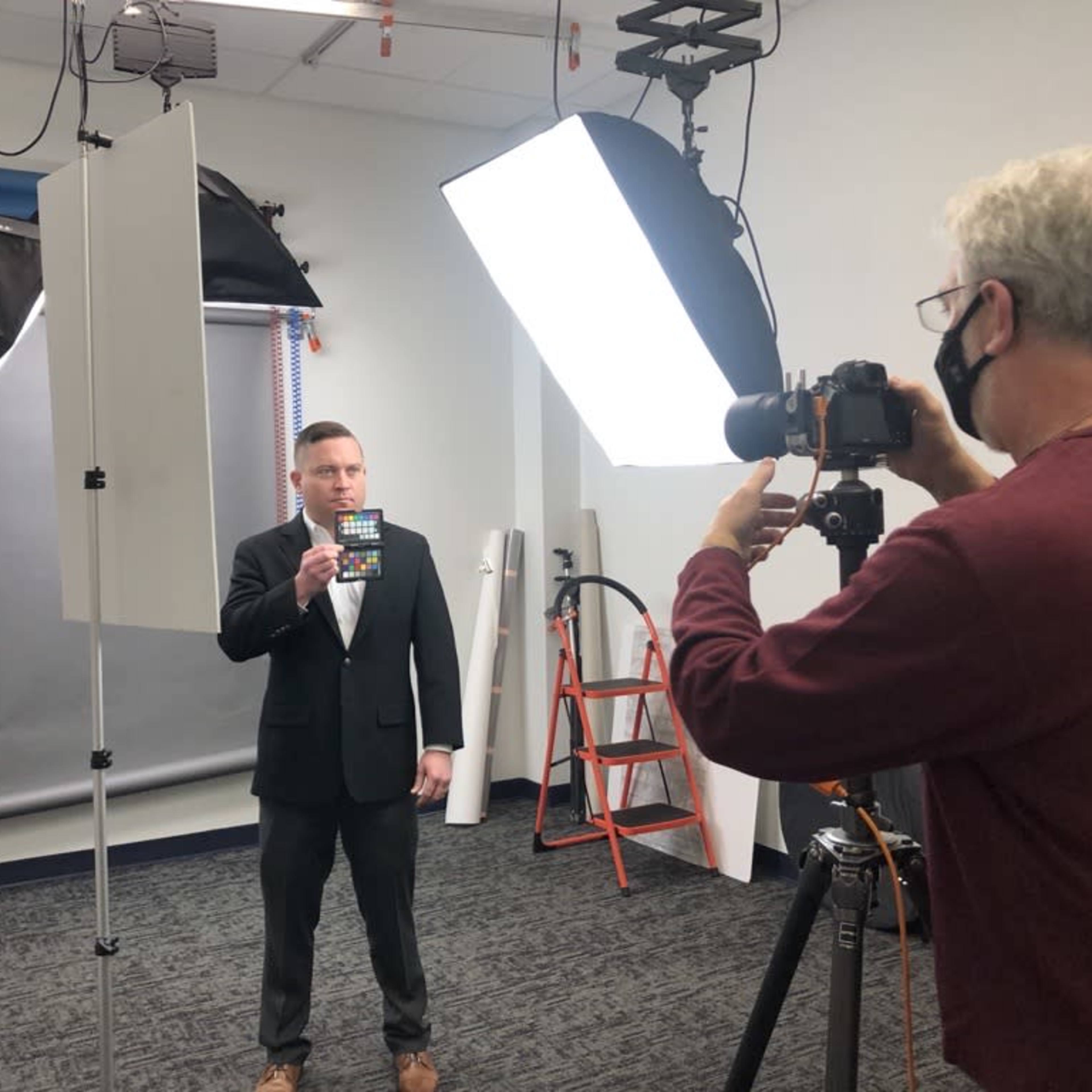 A man in a suit holds a slate while being photographed by another man in a studio setting with lighting equipment.