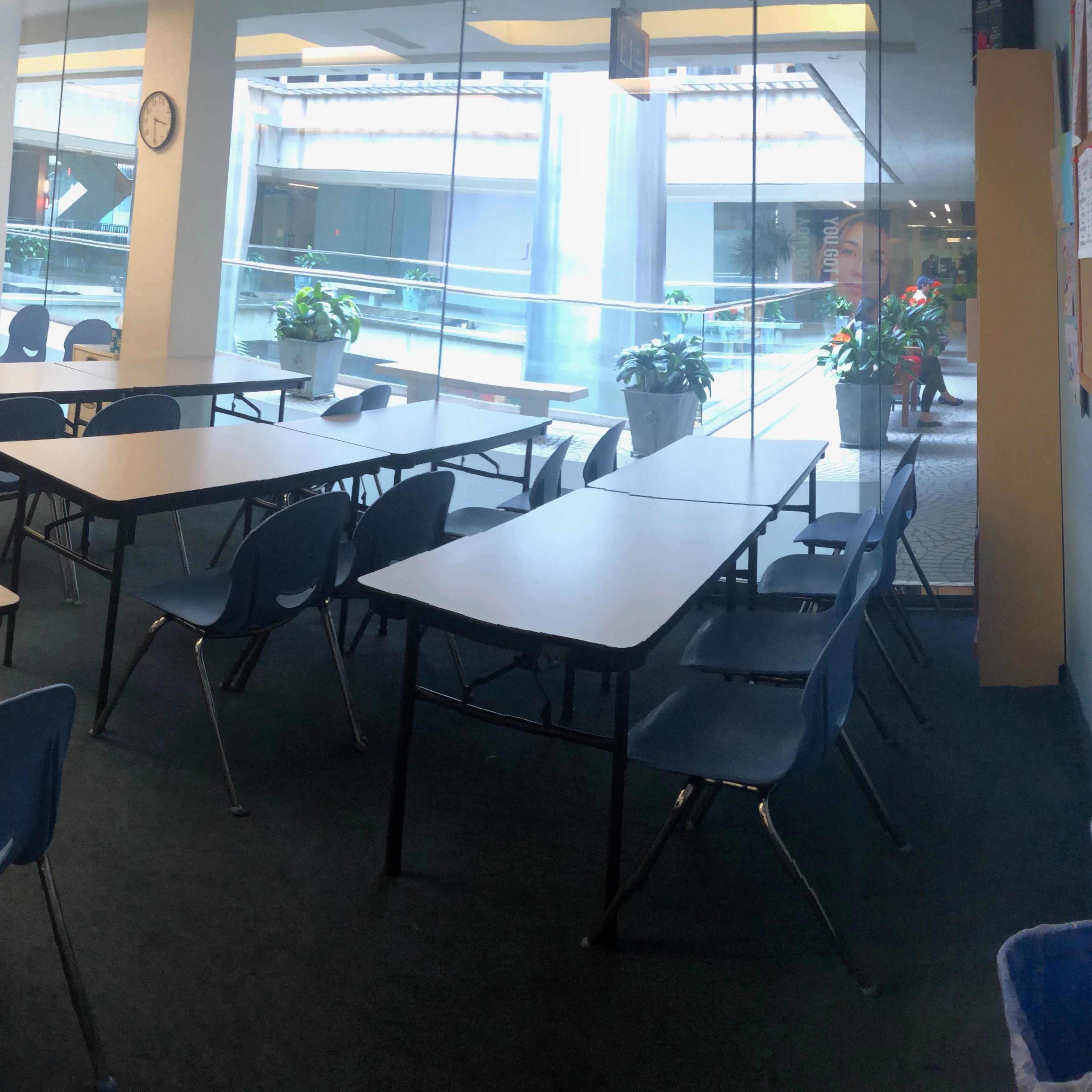 A classroom with several empty tables and chairs arranged in a circular formation, featuring large windows that provide a view of the outside.