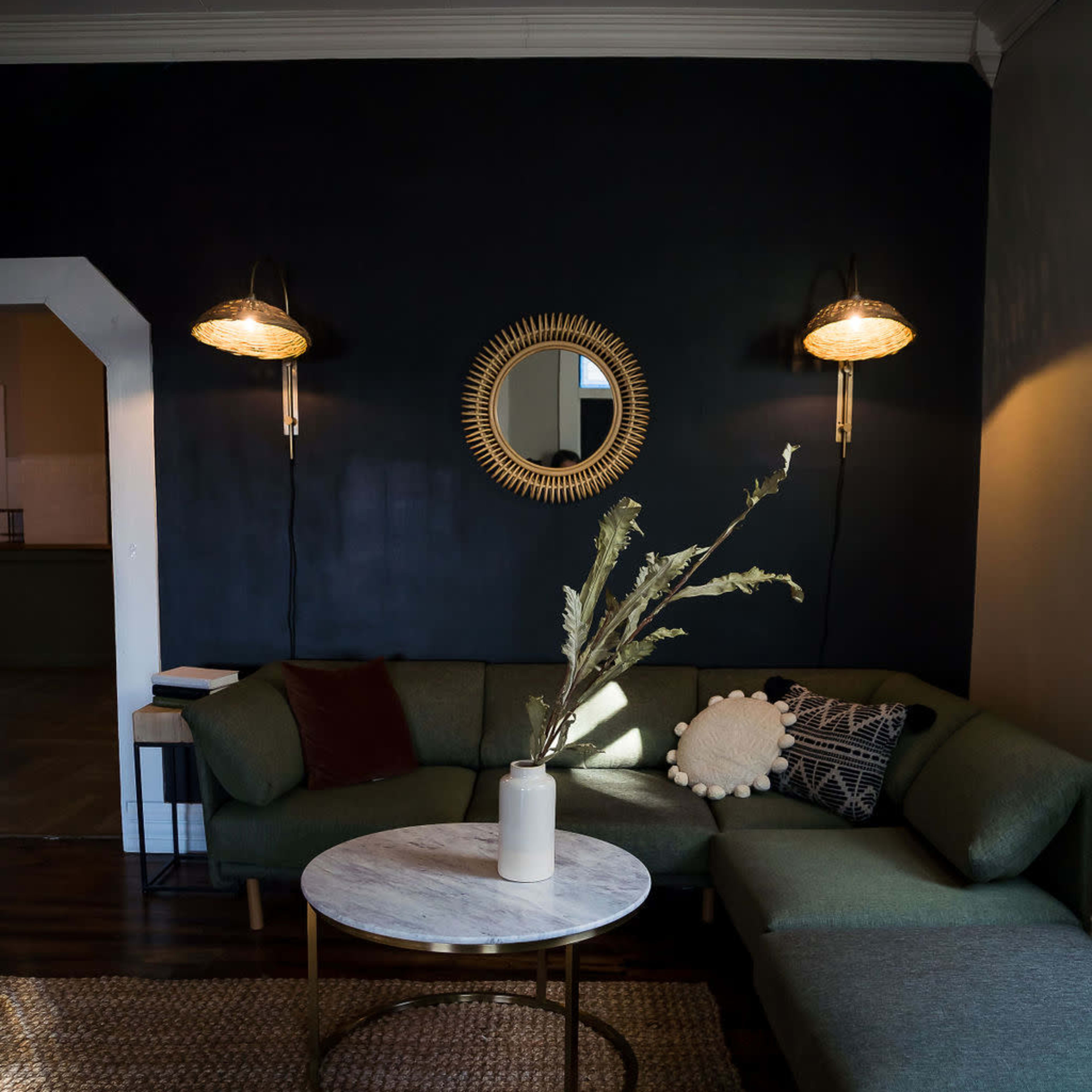 A cozy living room featuring a green sectional sofa, a round marble coffee table with a vase, and wall sconces illuminating a dark wall.