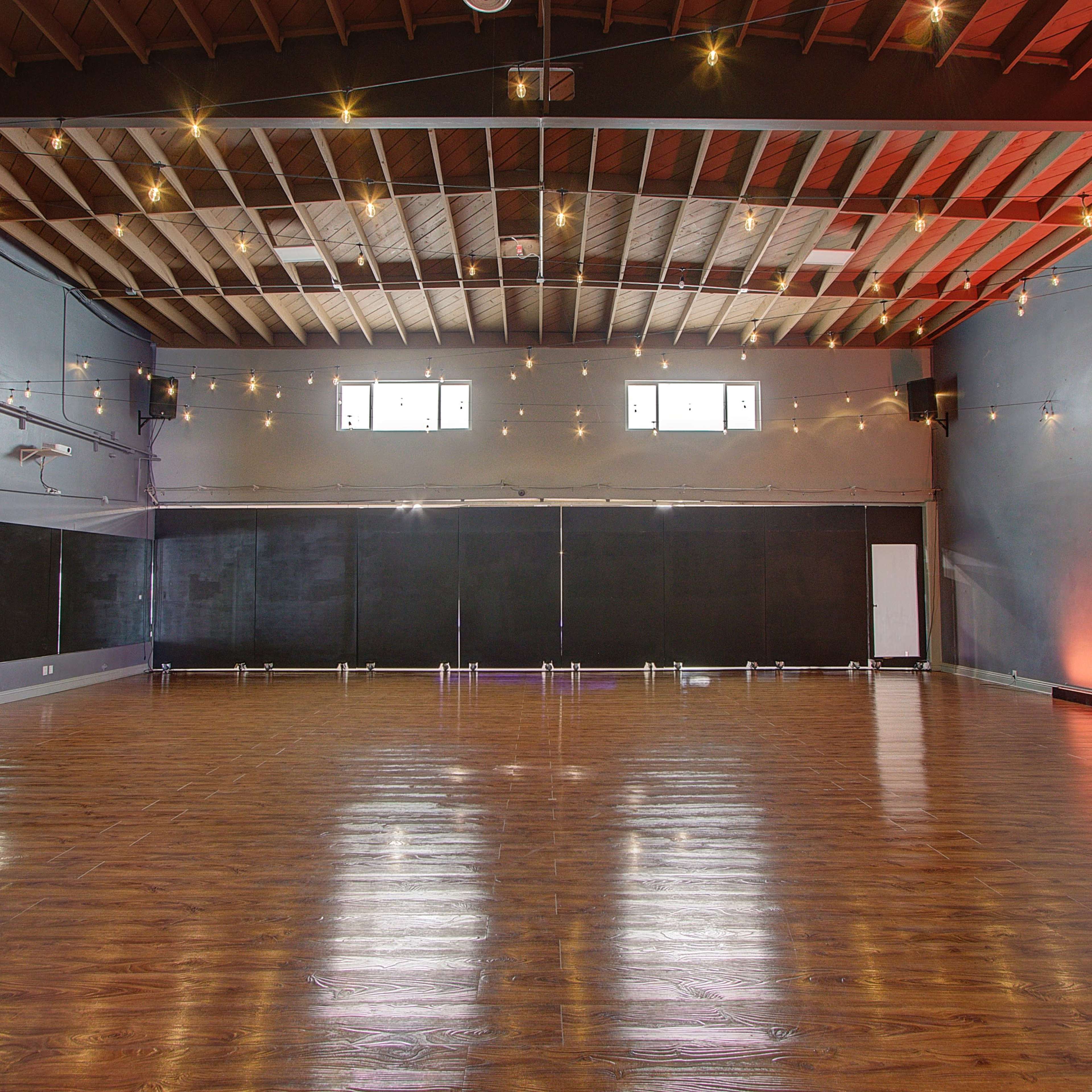 A spacious dance studio with polished wooden floors, mirrors along one wall, and string lights hanging from the ceiling.
