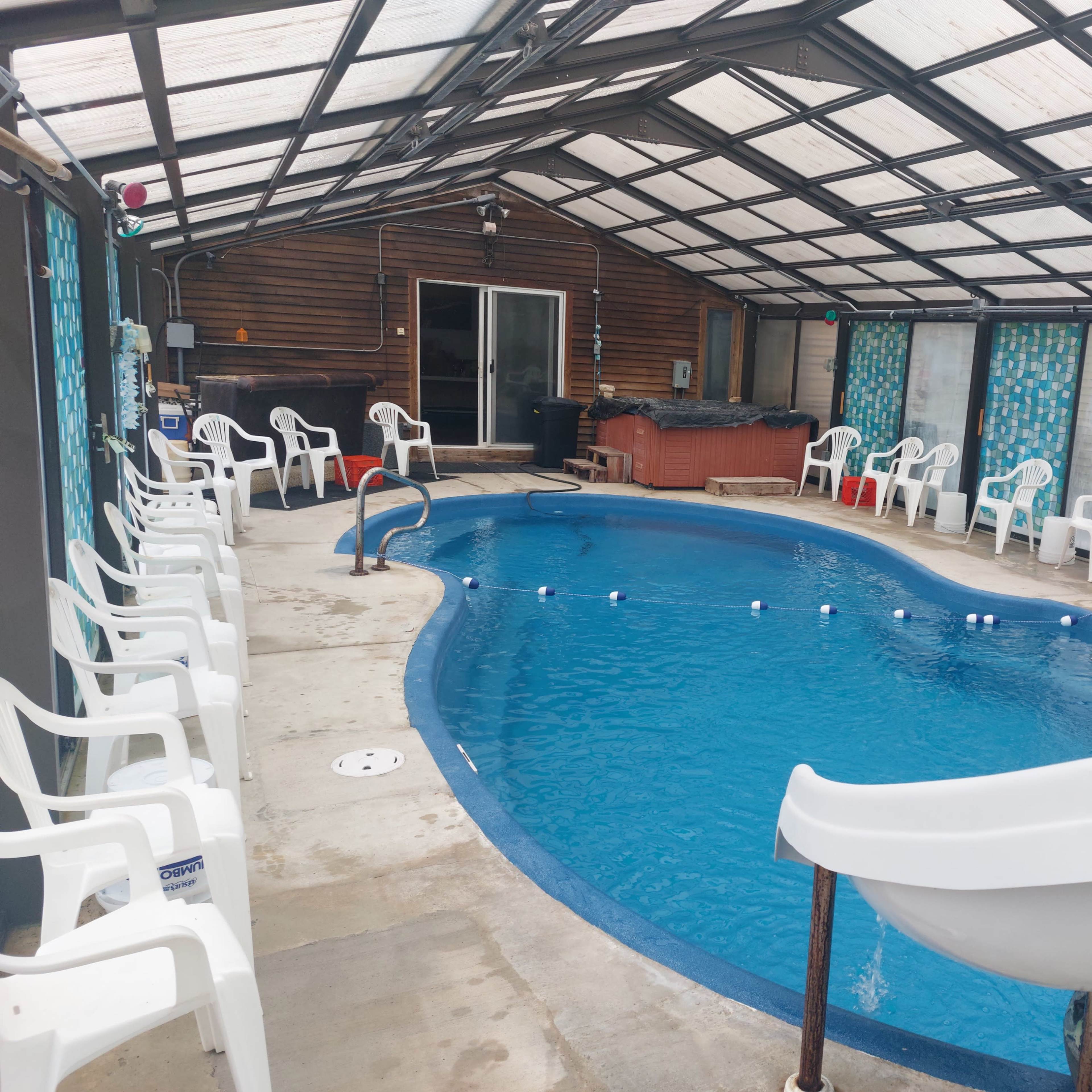 An indoor swimming pool area with a blue pool, a white waterslide, several white plastic chairs arranged around the perimeter, and a hot tub in the corner.