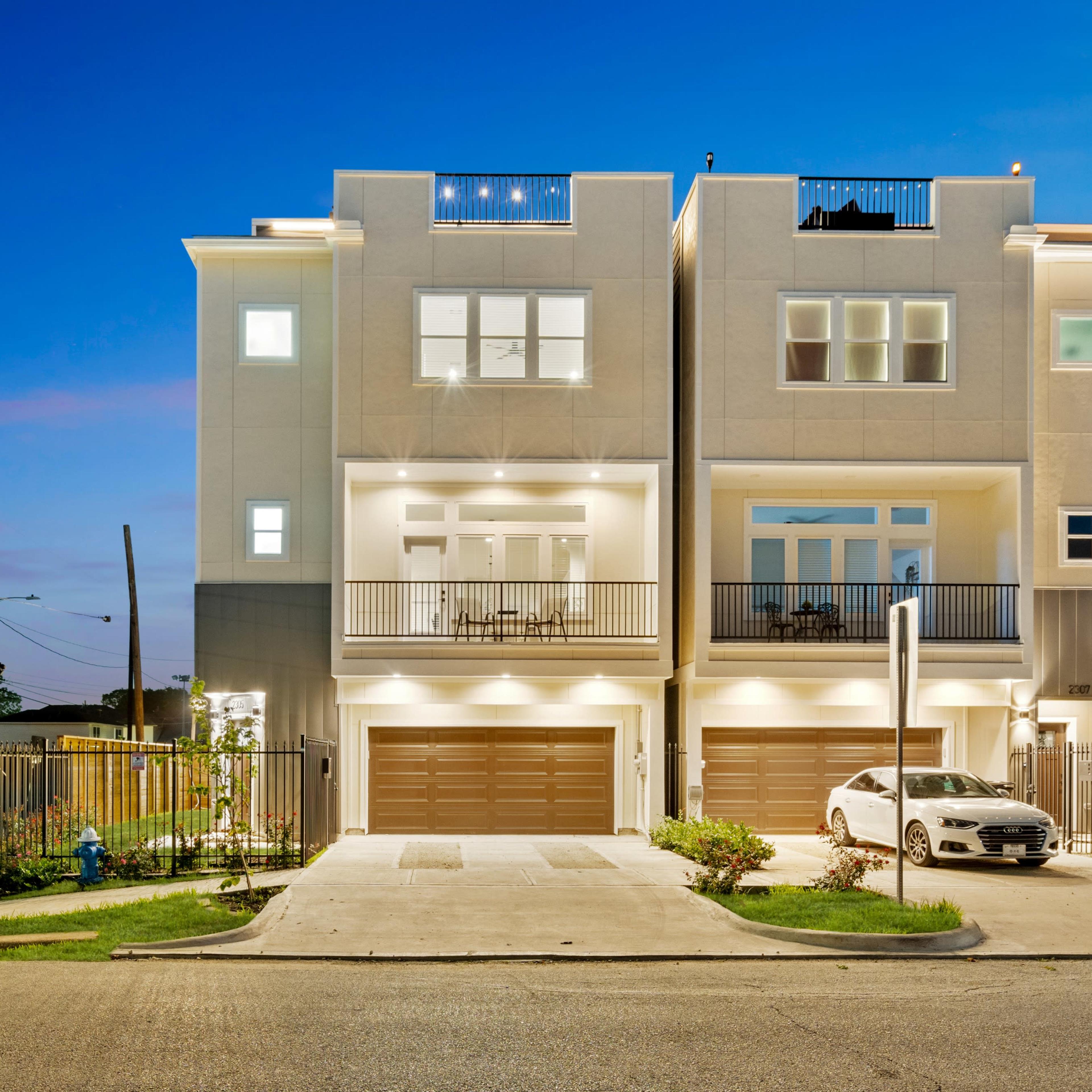 A modern townhouse with multiple levels, featuring three garage doors and balconies, illuminated at dusk.