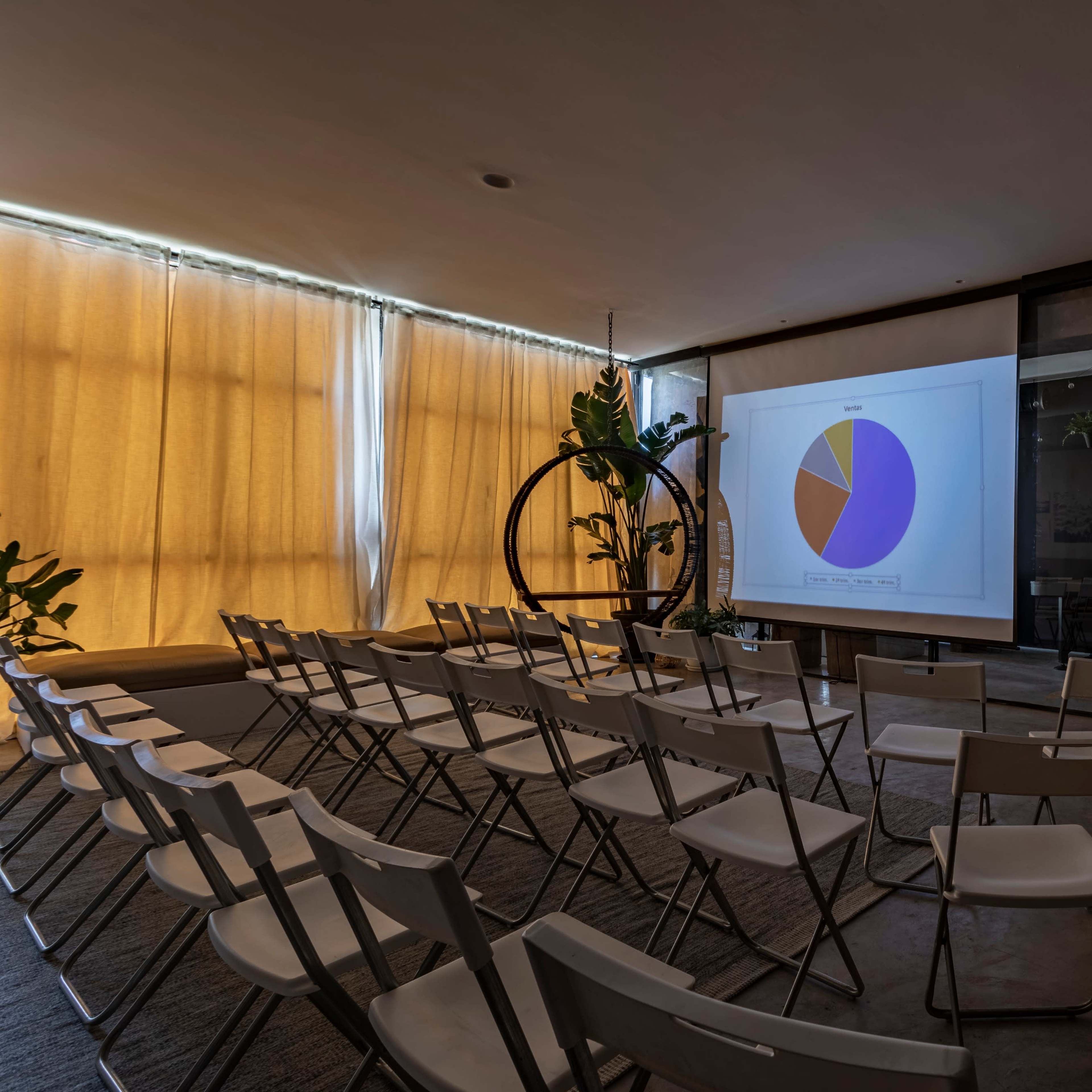 A presentation screen displaying a pie chart is set up in a sparsely furnished room with rows of folding chairs and potted plants.