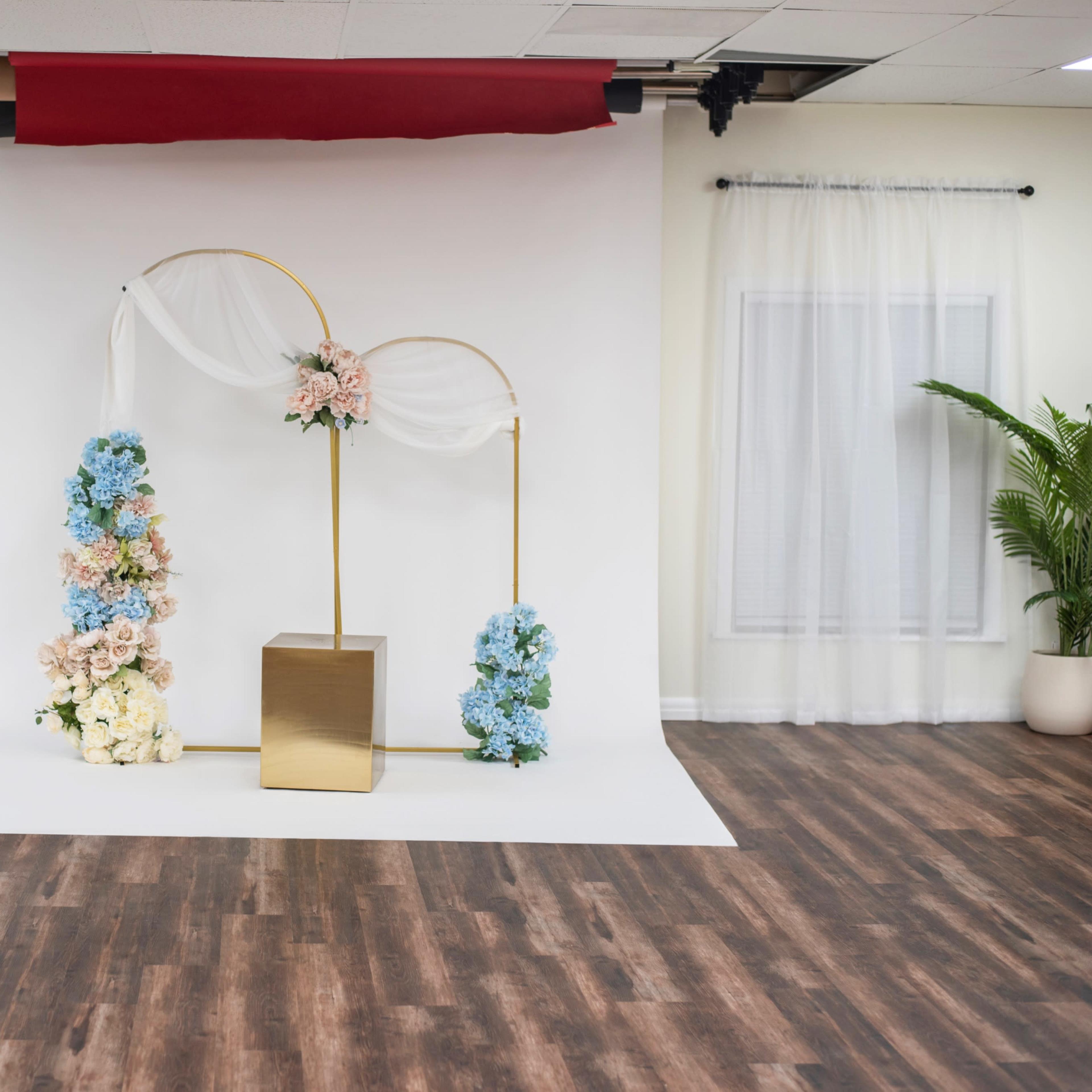 A decorated photo backdrop with floral arrangements on either side of a central gold structure in a room with light-colored walls and wood-like flooring.
