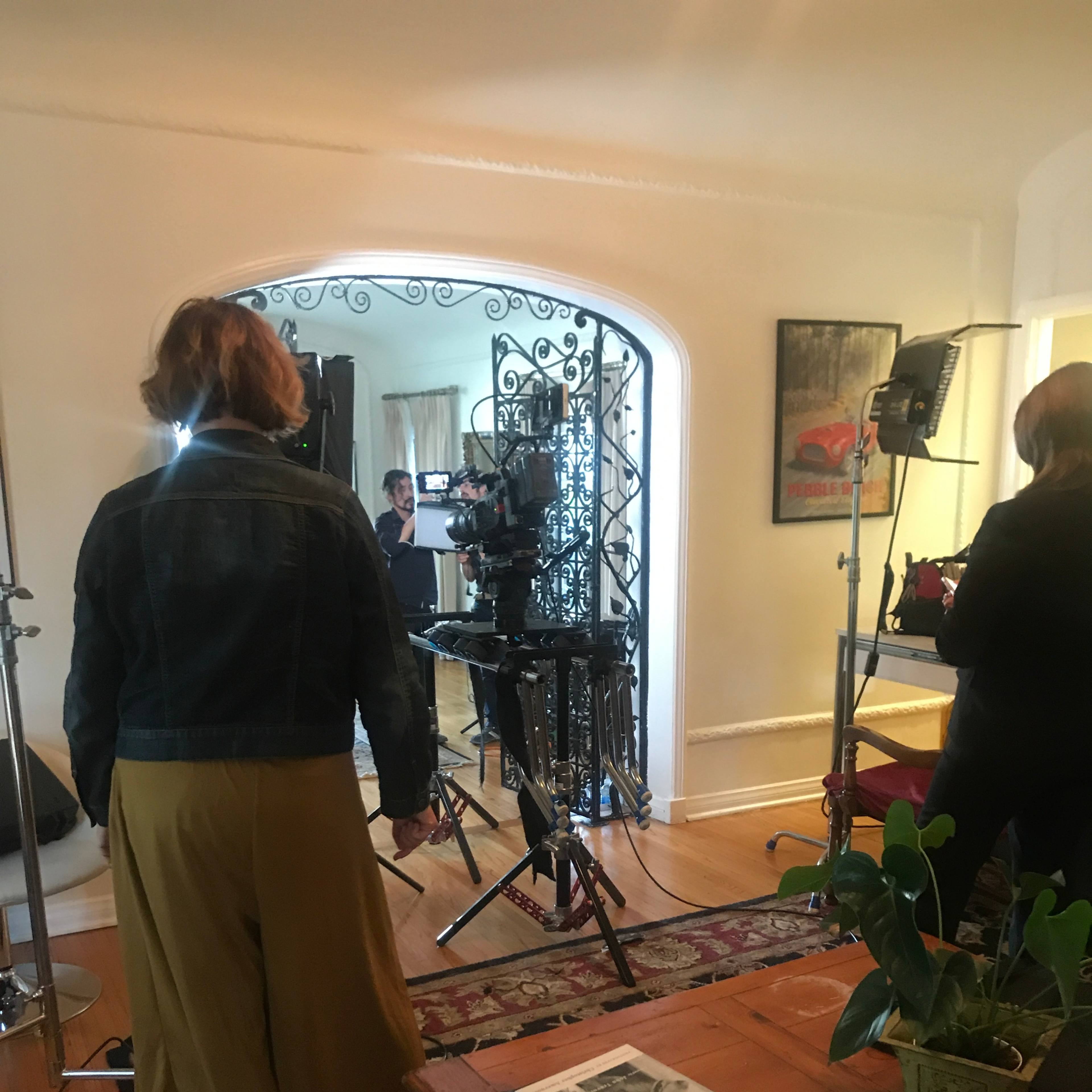 A film crew sets up equipment in a living room, preparing to shoot a scene with cameras and lights.