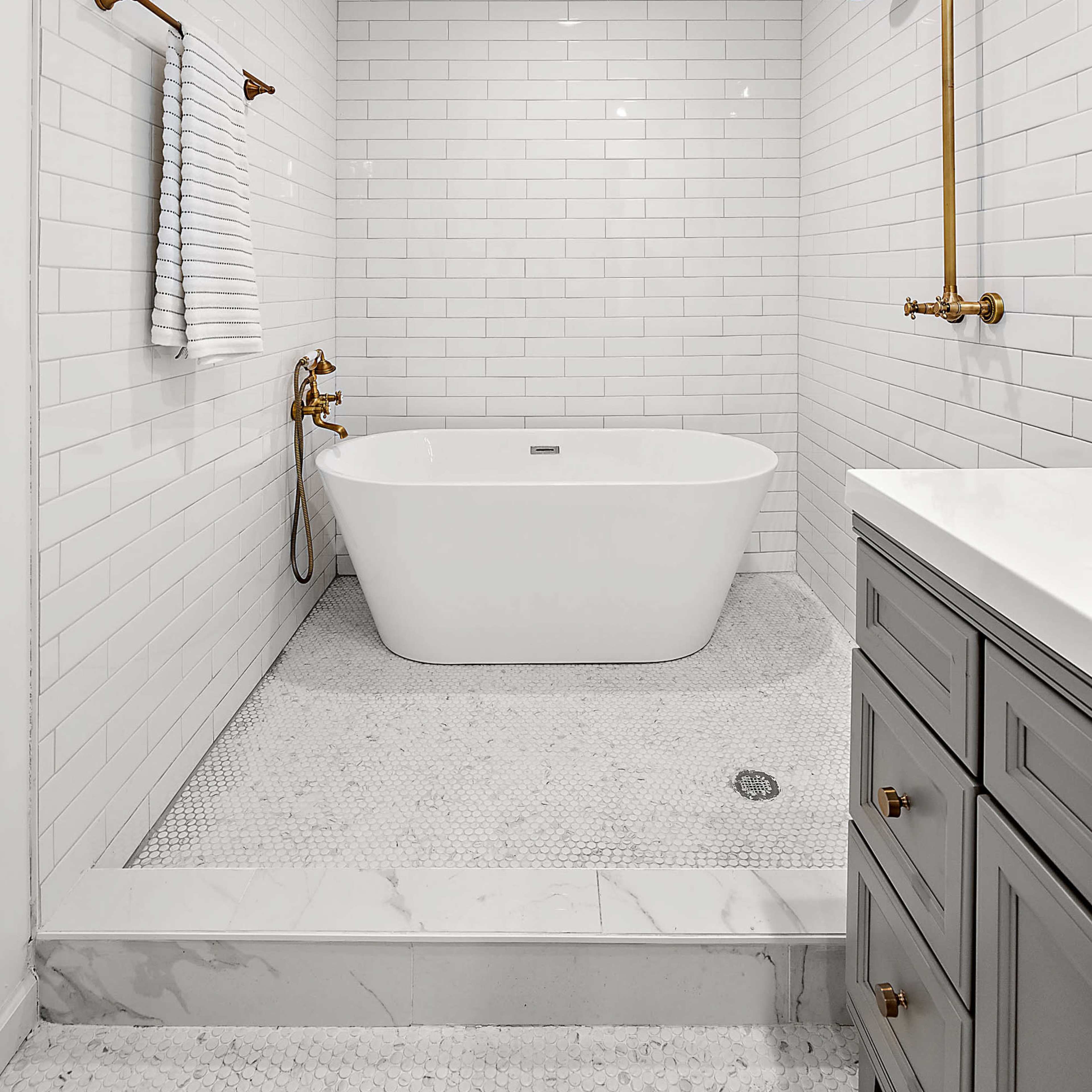 The bathroom features a freestanding white tub, a rainfall showerhead, and a sink with gray cabinetry, all set against a backdrop of white subway tile and a marble floor.