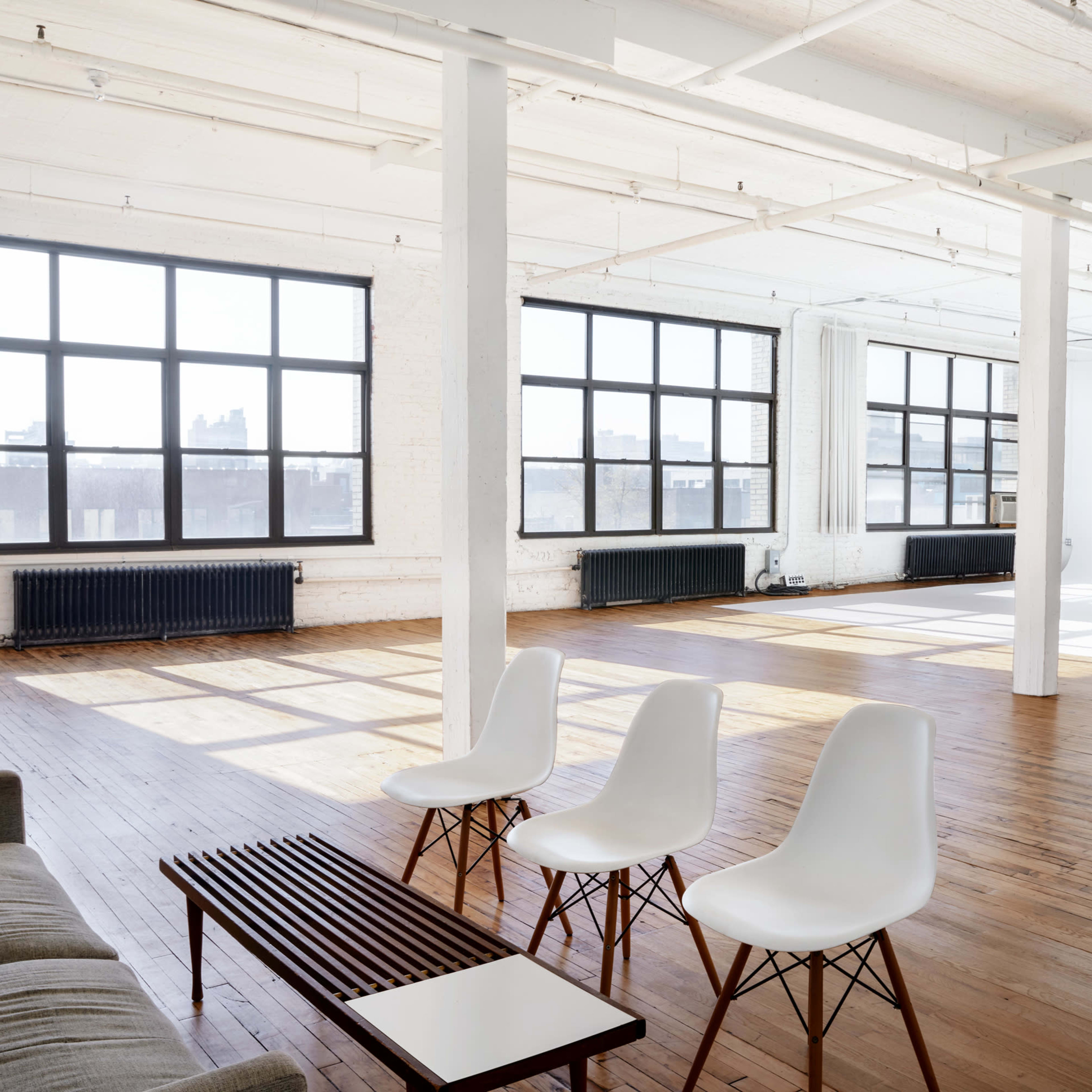 An airy, open space features large windows, wooden floors, a sofa, and three white chairs arranged around a low table.
