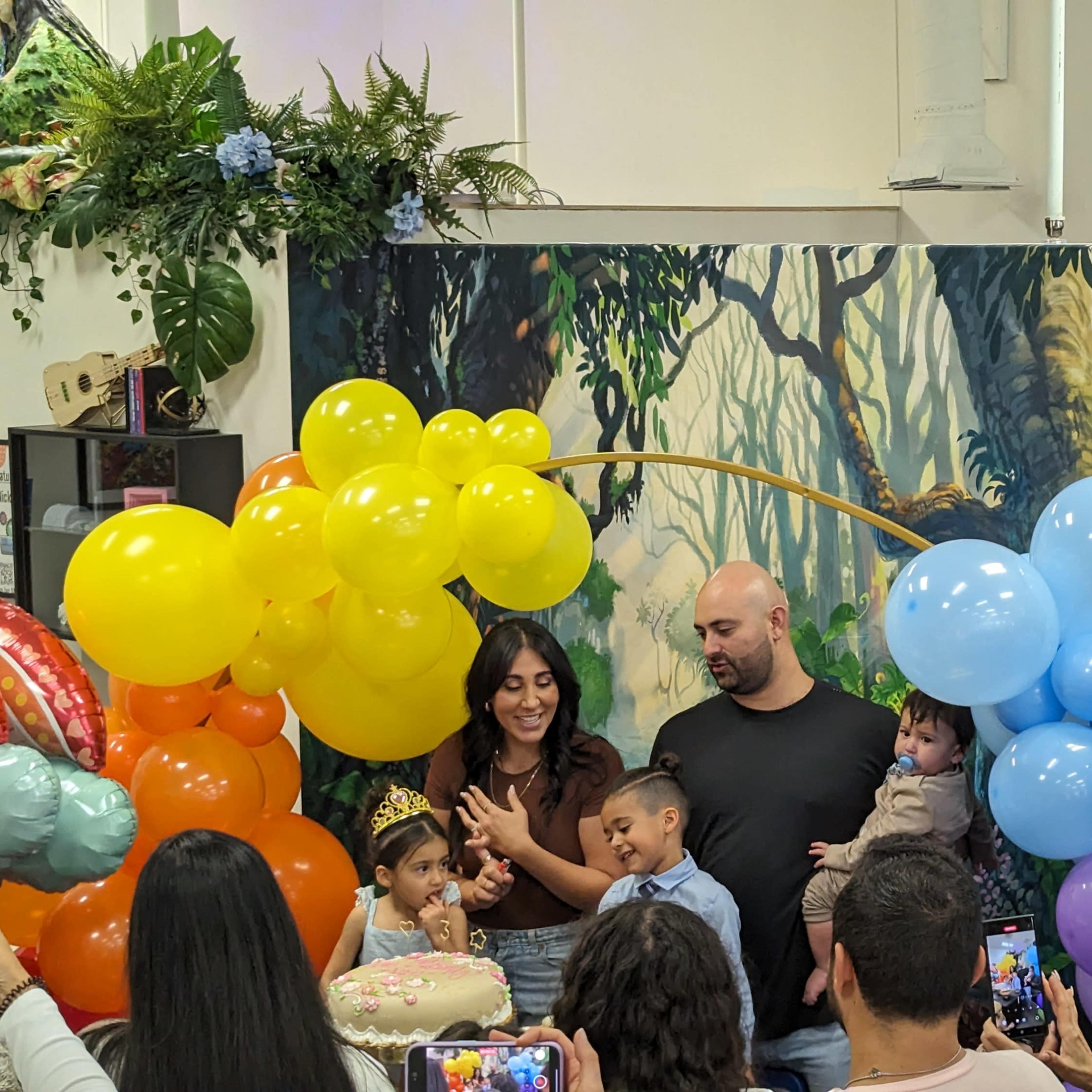 A family gathers around a decorated birthday cake amidst colorful balloons, while attendees capture the moment with their phones.