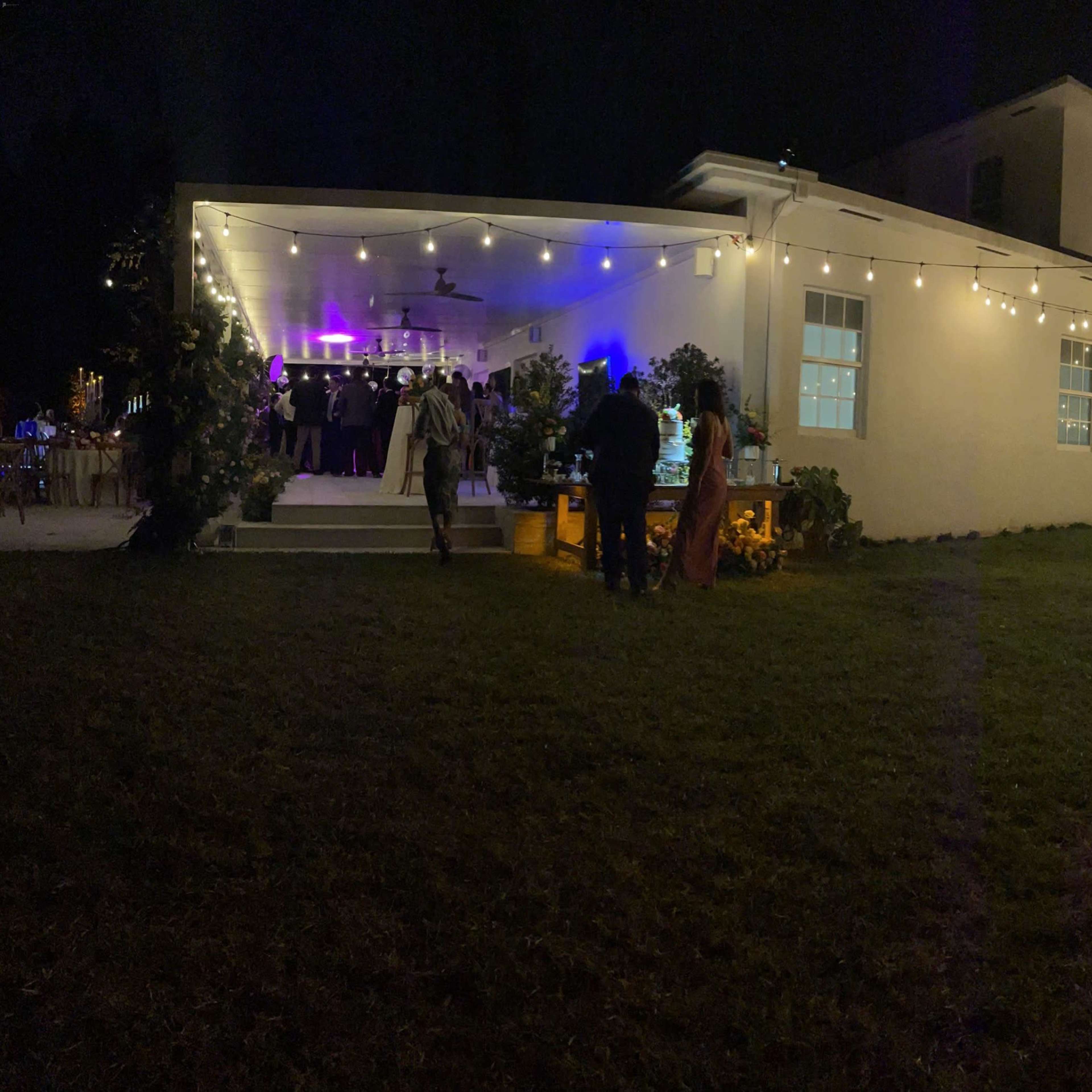 A large outdoor gathering takes place near a well-lit house adorned with string lights and people socializing in the yard.