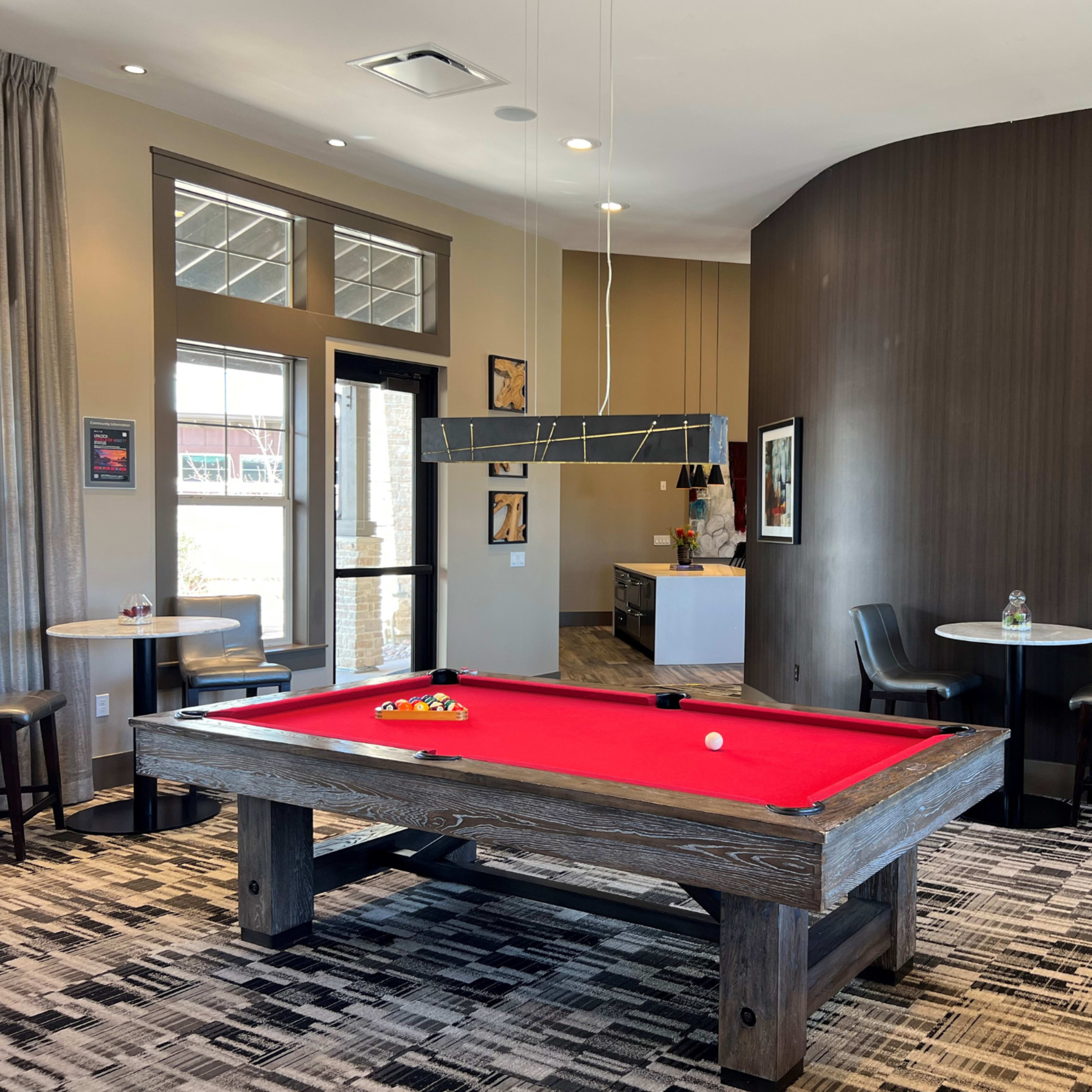 A game room featuring a red-felt pool table, two small tables with chairs, and a wall-mounted television.
