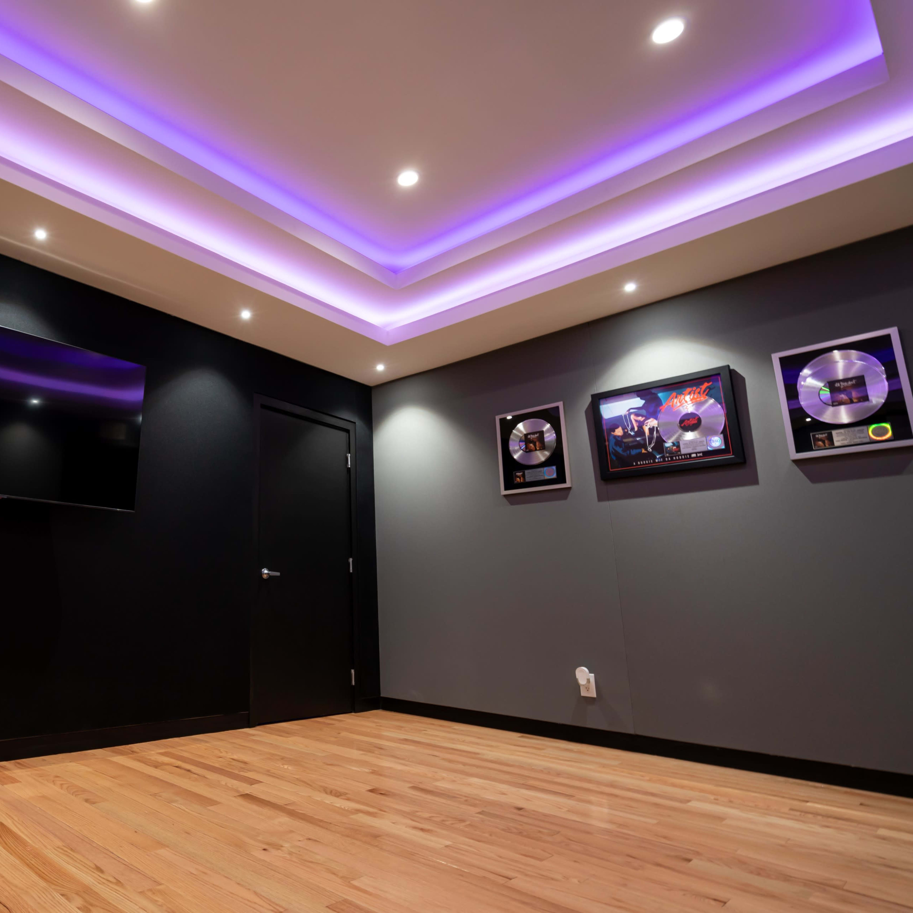 A music studio with a dark wall, wooden flooring, a flat-screen TV mounted on one wall, and framed awards hanging on another wall under a purple-lit ceiling.