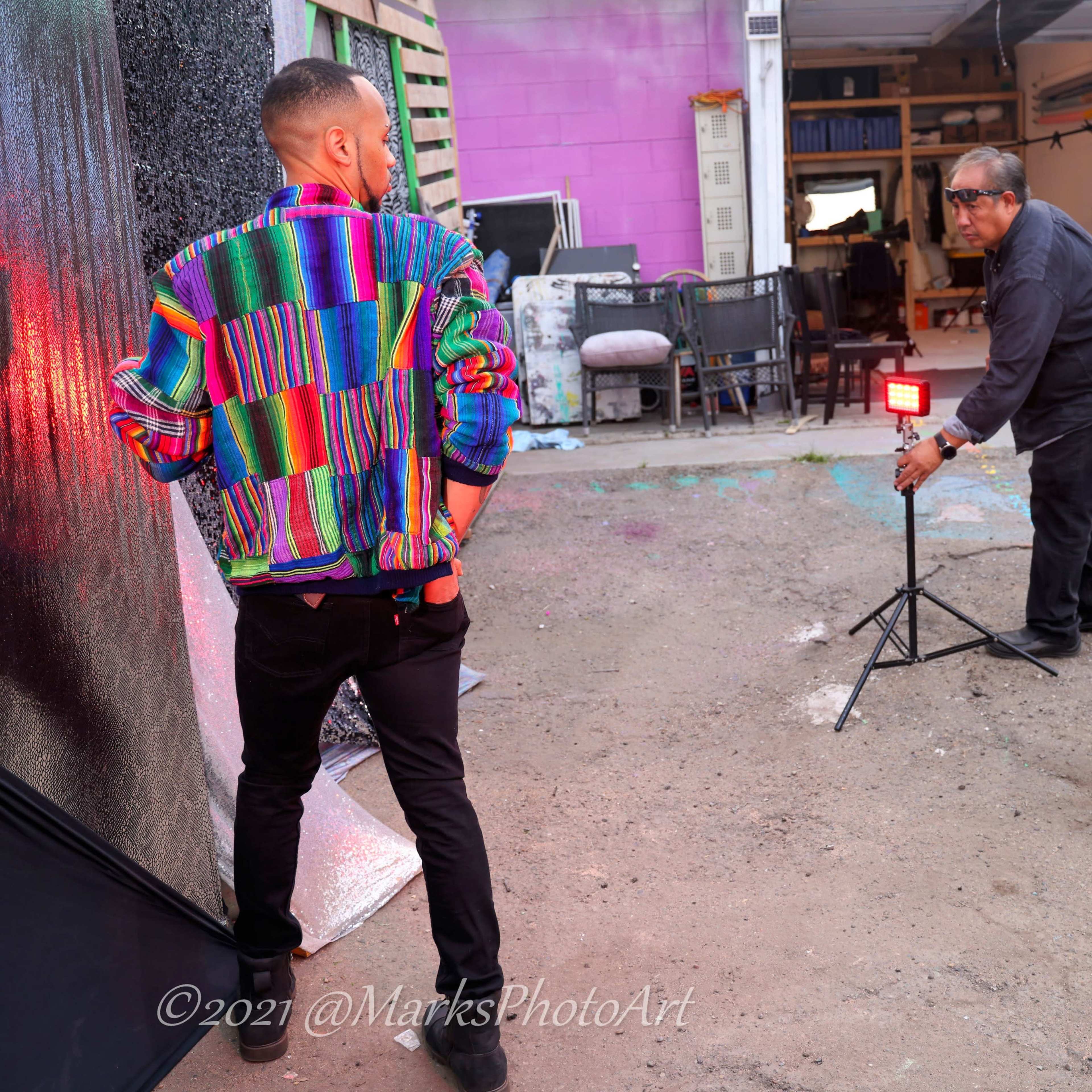A man poses in a colorful jacket while two photographers set up lighting equipment for a photoshoot in an outdoor space.