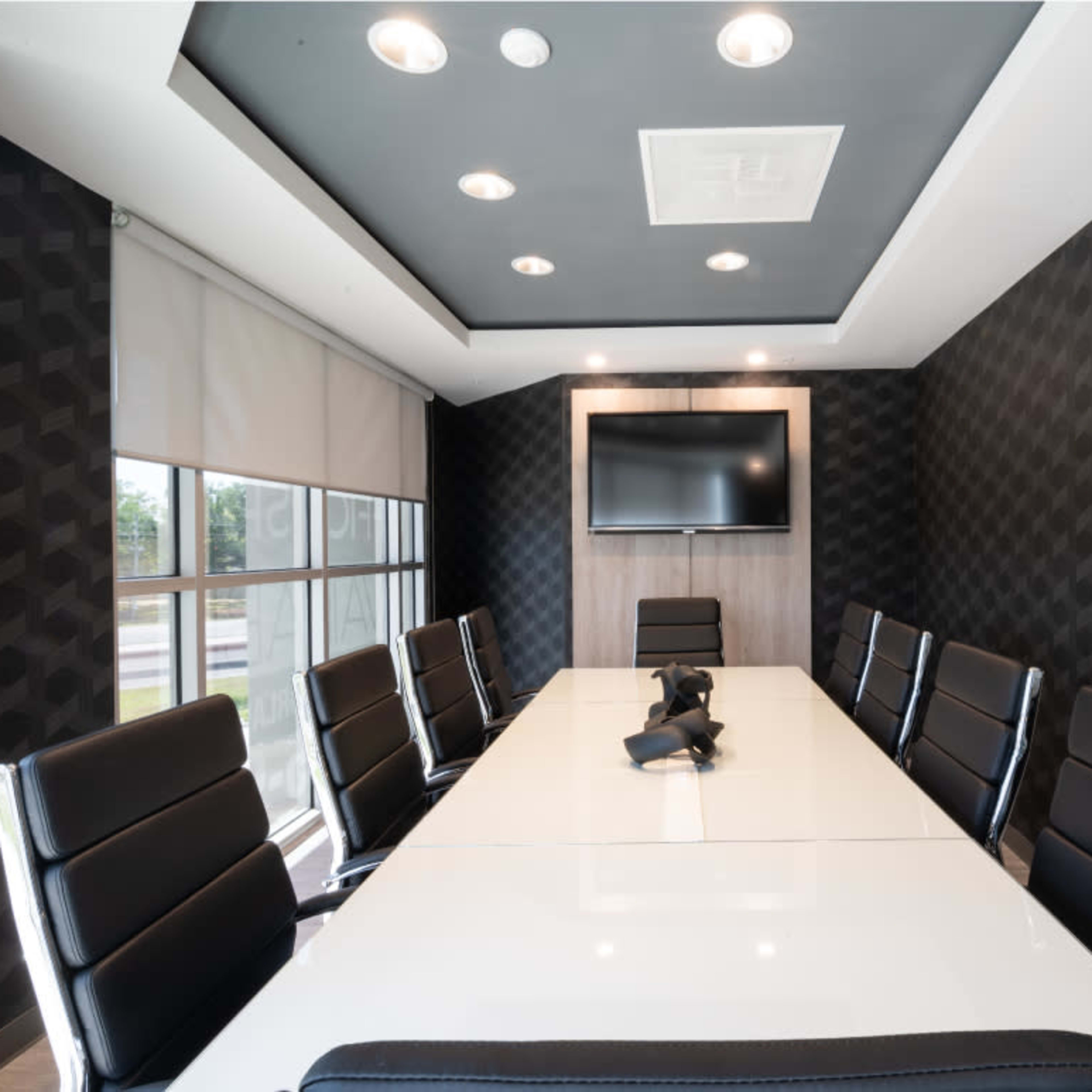 A modern conference room features a long white table surrounded by black chairs, with a wall-mounted television and geometric patterned wallpaper.