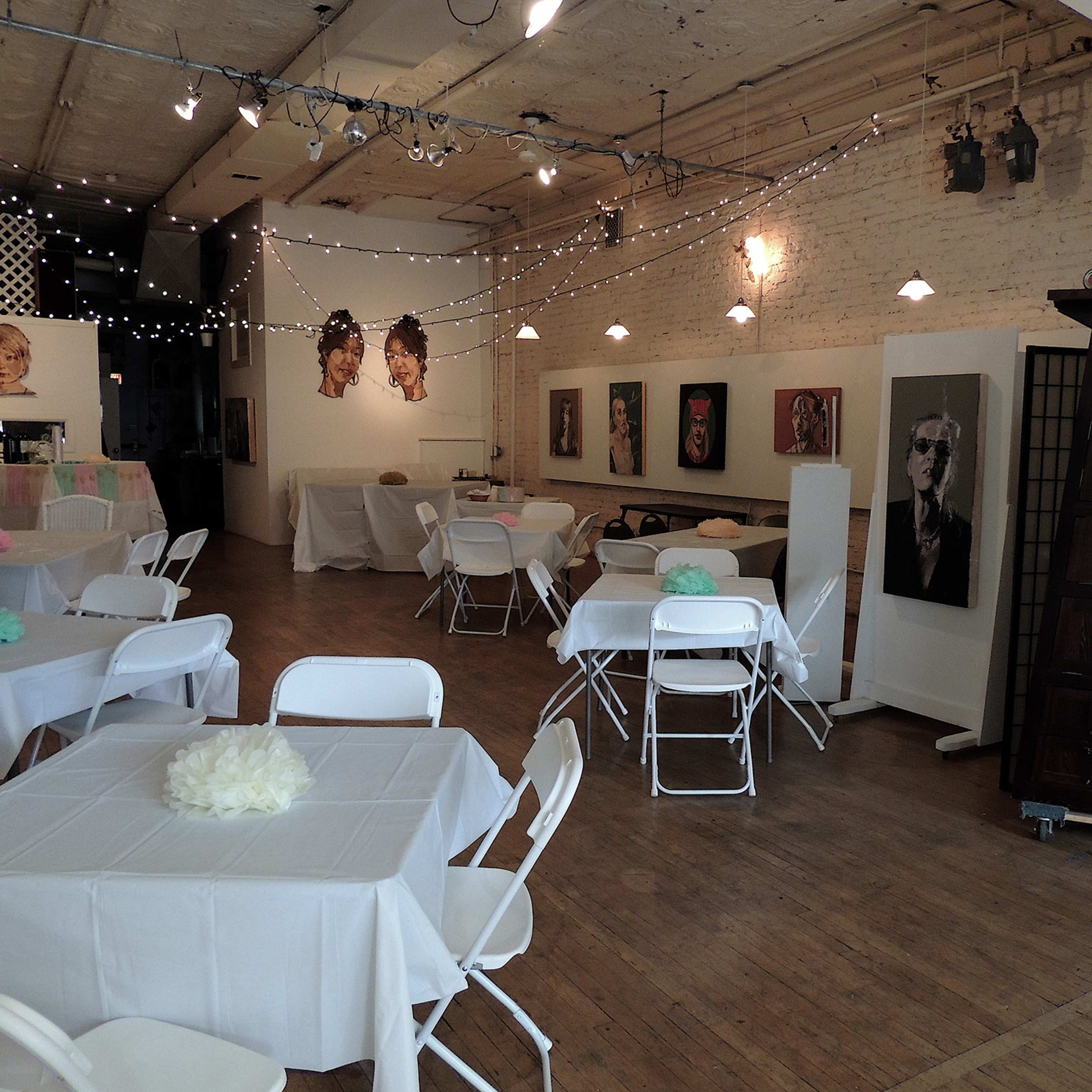 An event space with white tables and chairs, decorated with light-colored floral centerpieces, and featuring art on the walls and string lights overhead.
