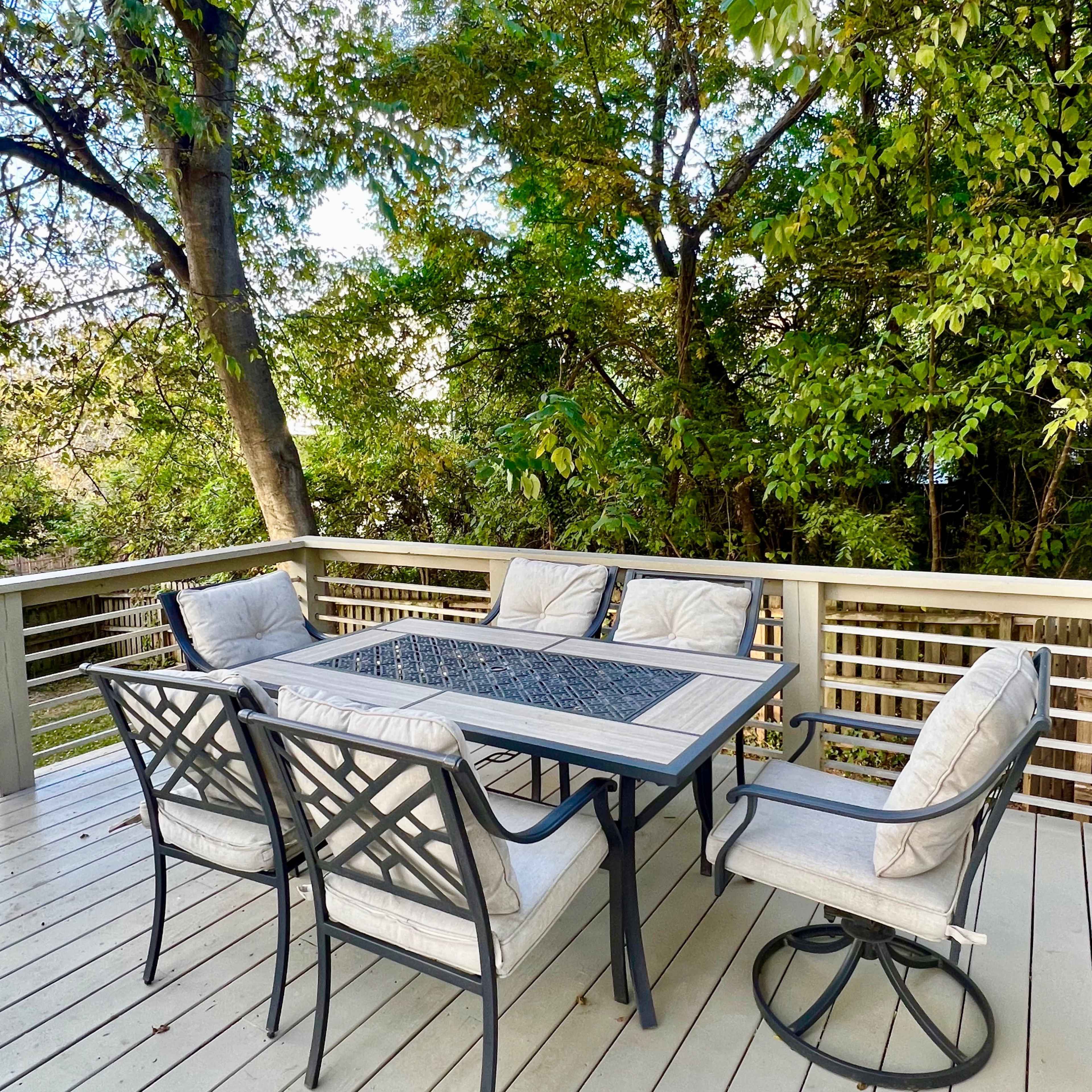 A patio dining set with a rectangular table is surrounded by four cushioned chairs on a wooden deck amid lush greenery.