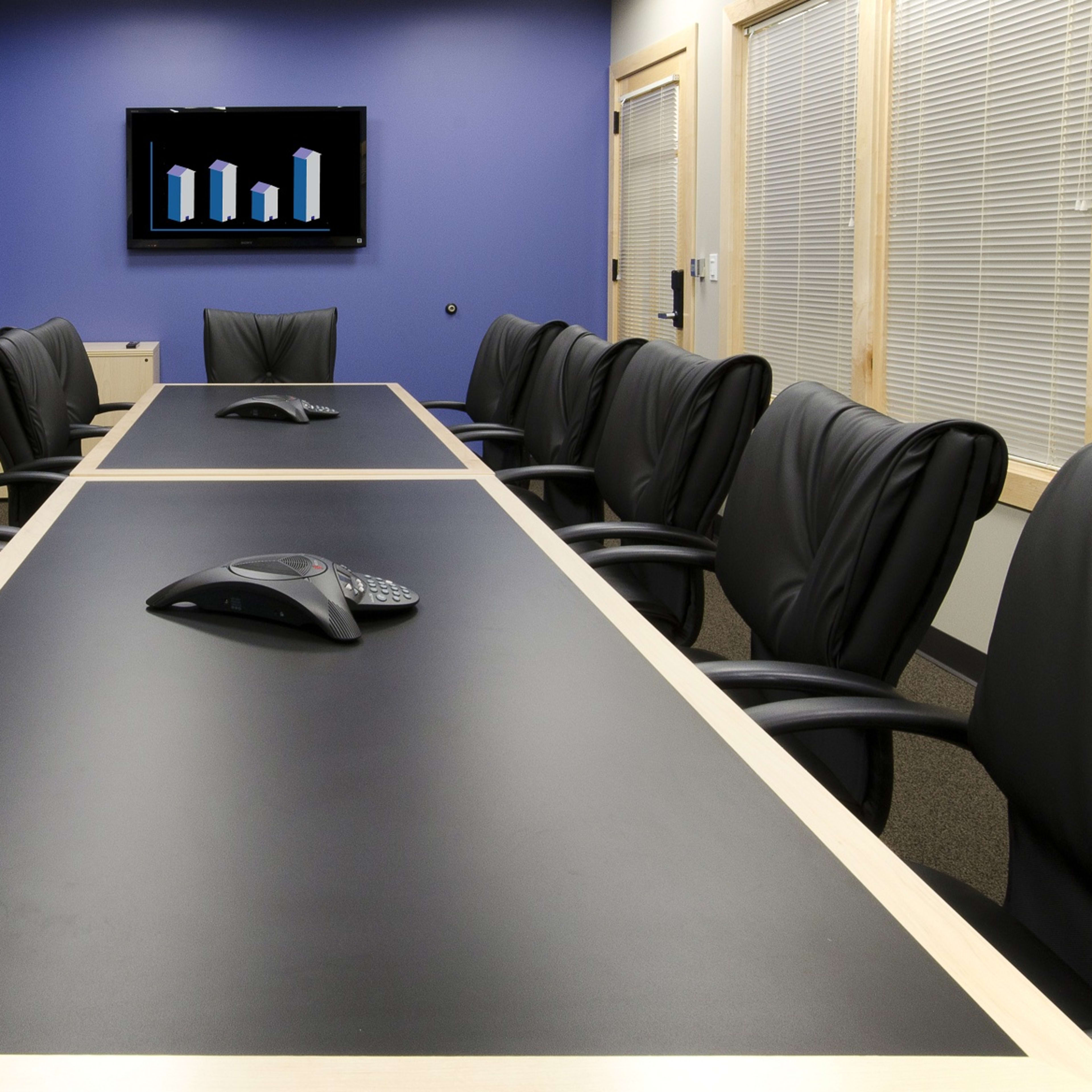 A conference room features a long black table with several black leather chairs, a video conference device in the center, and a wall-mounted screen displaying bar graphs.