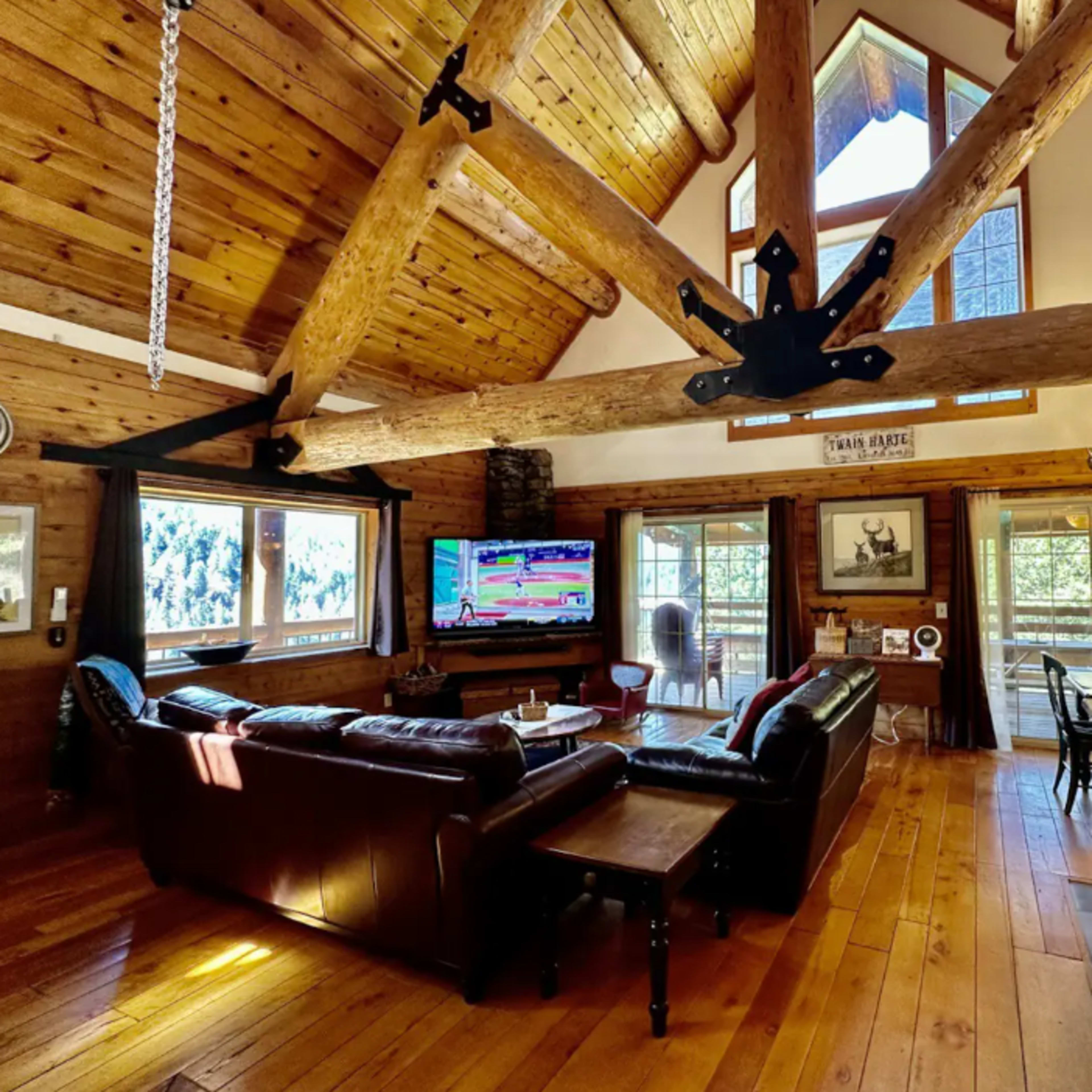 A spacious log cabin living room featuring a large leather sofa, a television, and large windows that offer views of the surrounding nature.