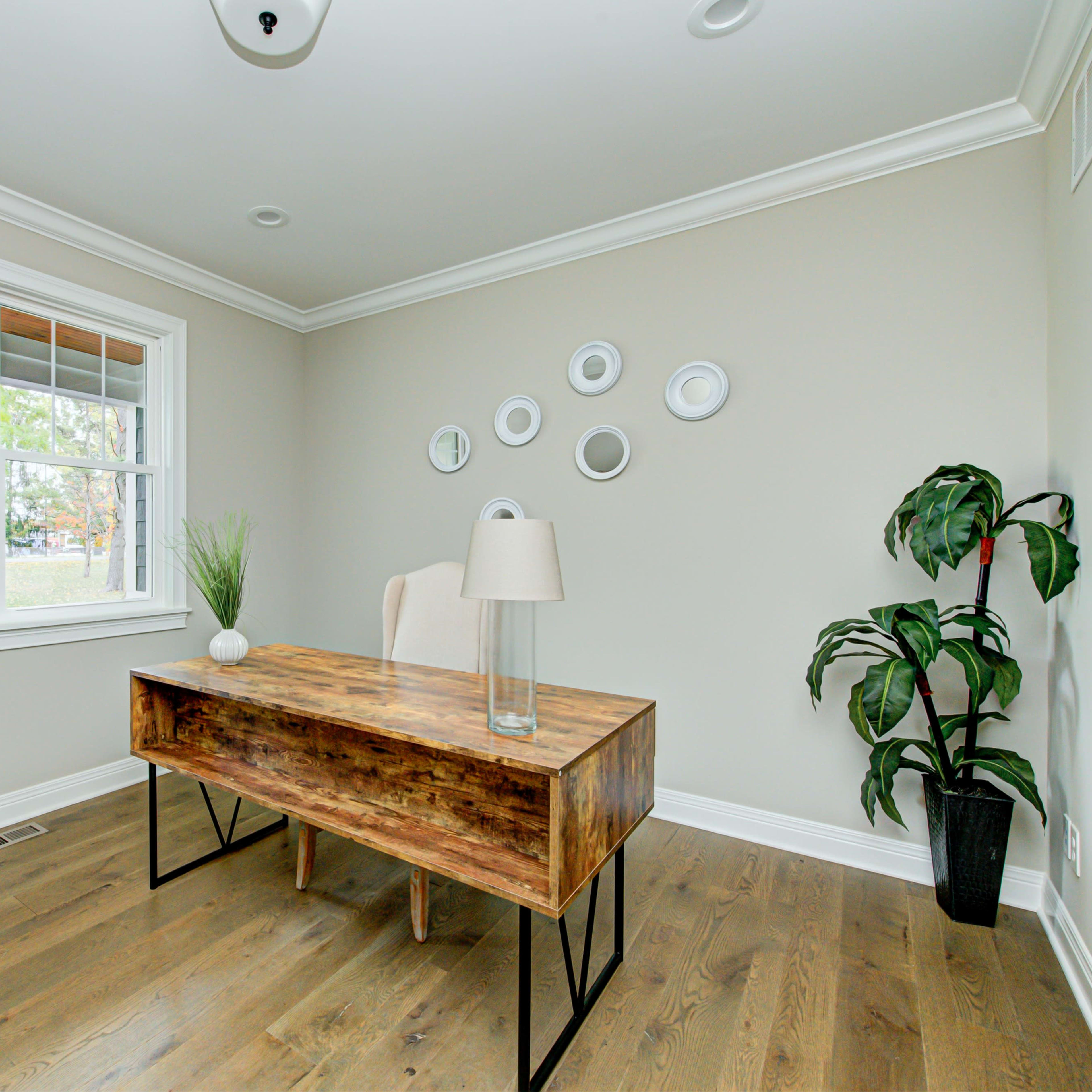 A modern office space featuring a wooden desk, a lamp, a chair, a potted plant, and decorative wall circles, with a window allowing natural light to enter.