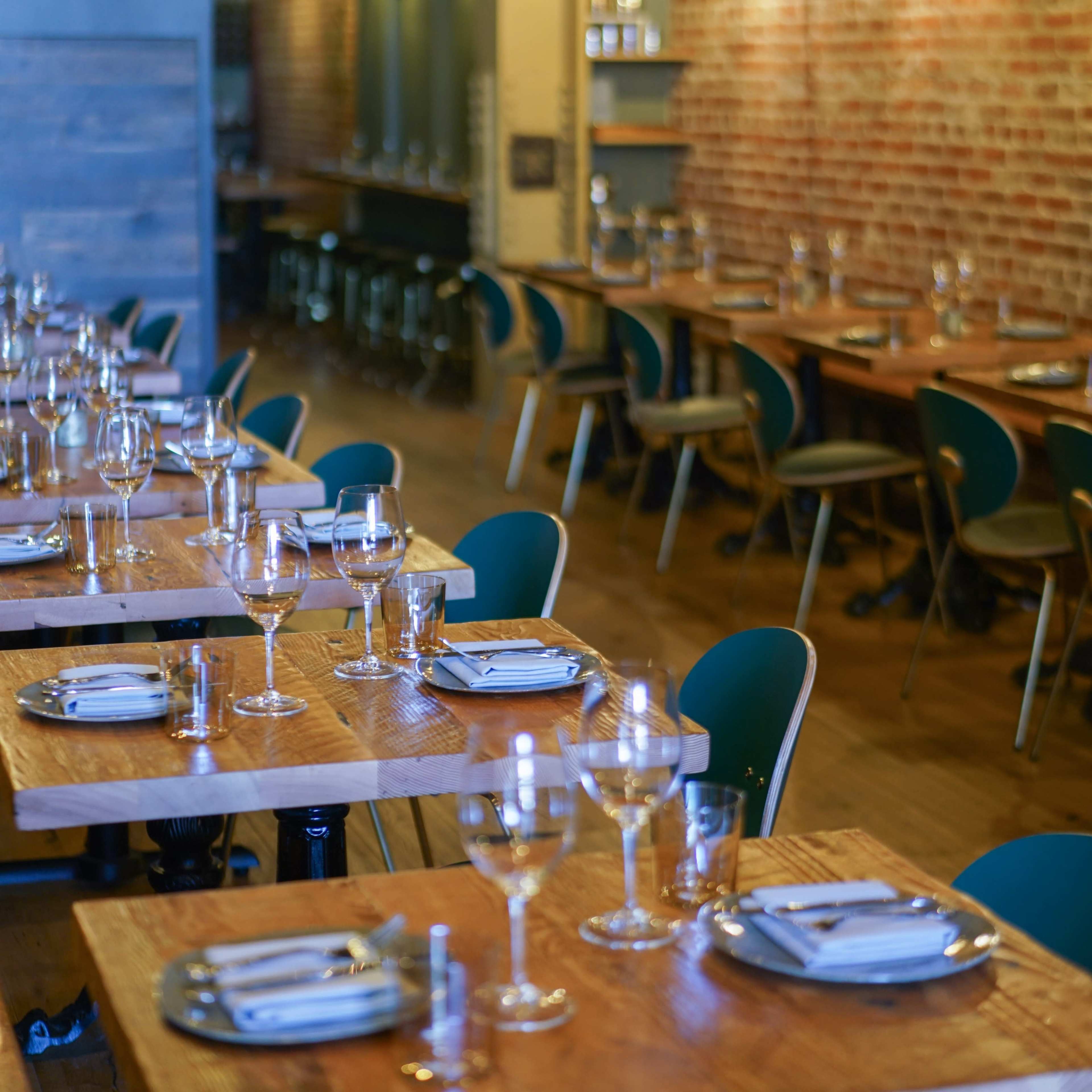 A restaurant interior with long, wooden tables set for dining, featuring glassware and cutlery, against a backdrop of exposed brick walls and minimalist decor.
