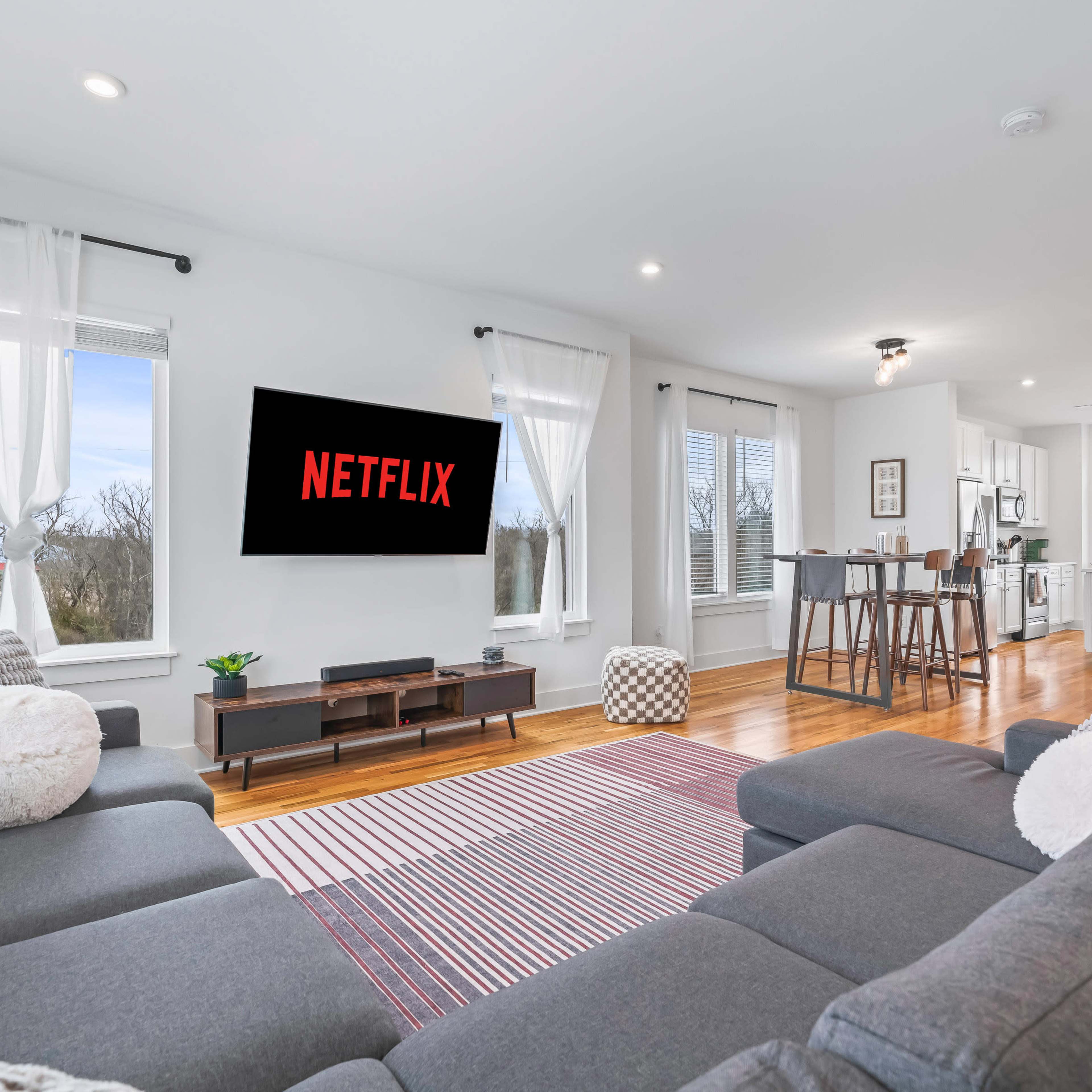 A modern living room features a large sectional sofa, a wall-mounted TV displaying the Netflix logo, and an open kitchen in the background.