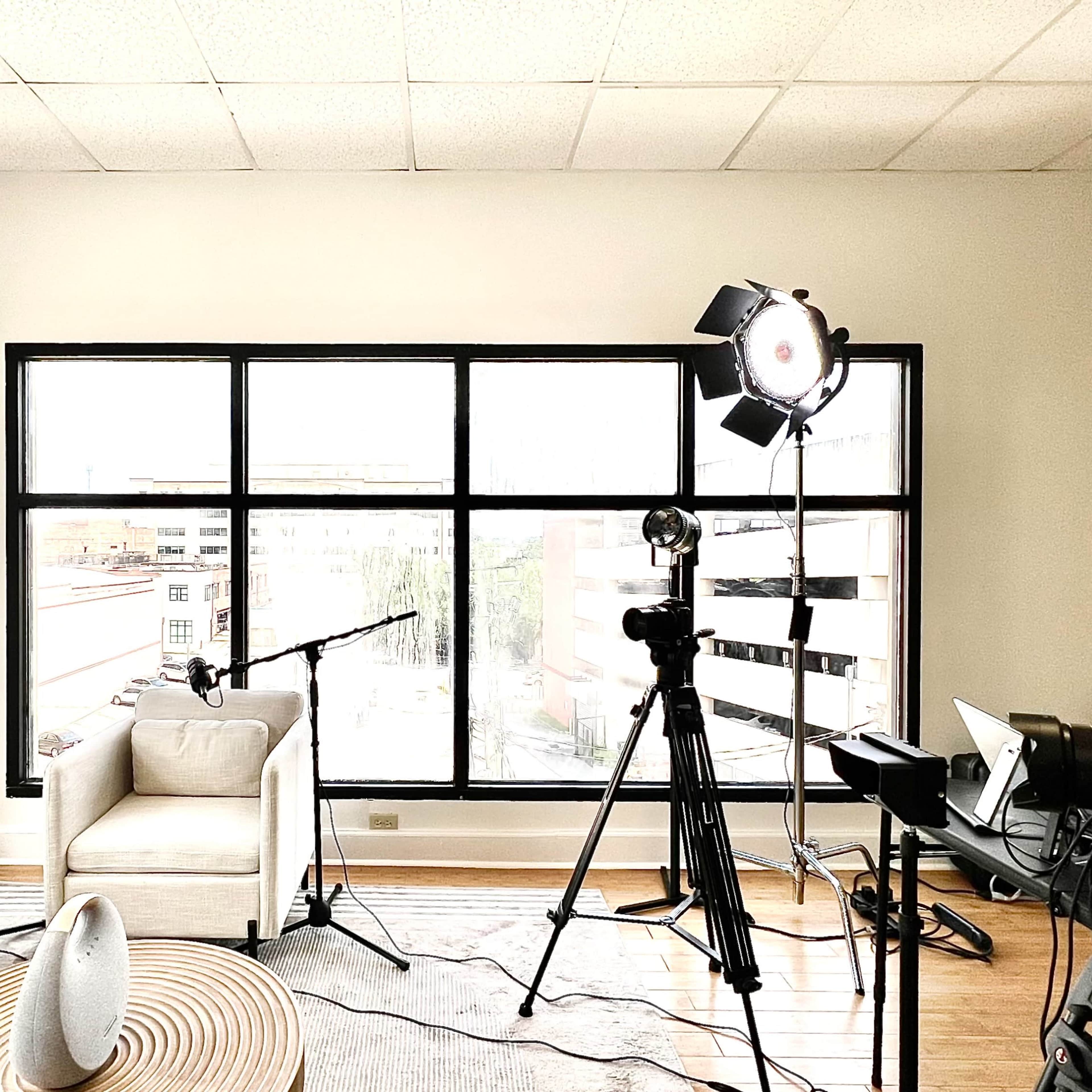 A film or photo studio setup featuring two chairs, a large window, lighting equipment, and a camera on a tripod.